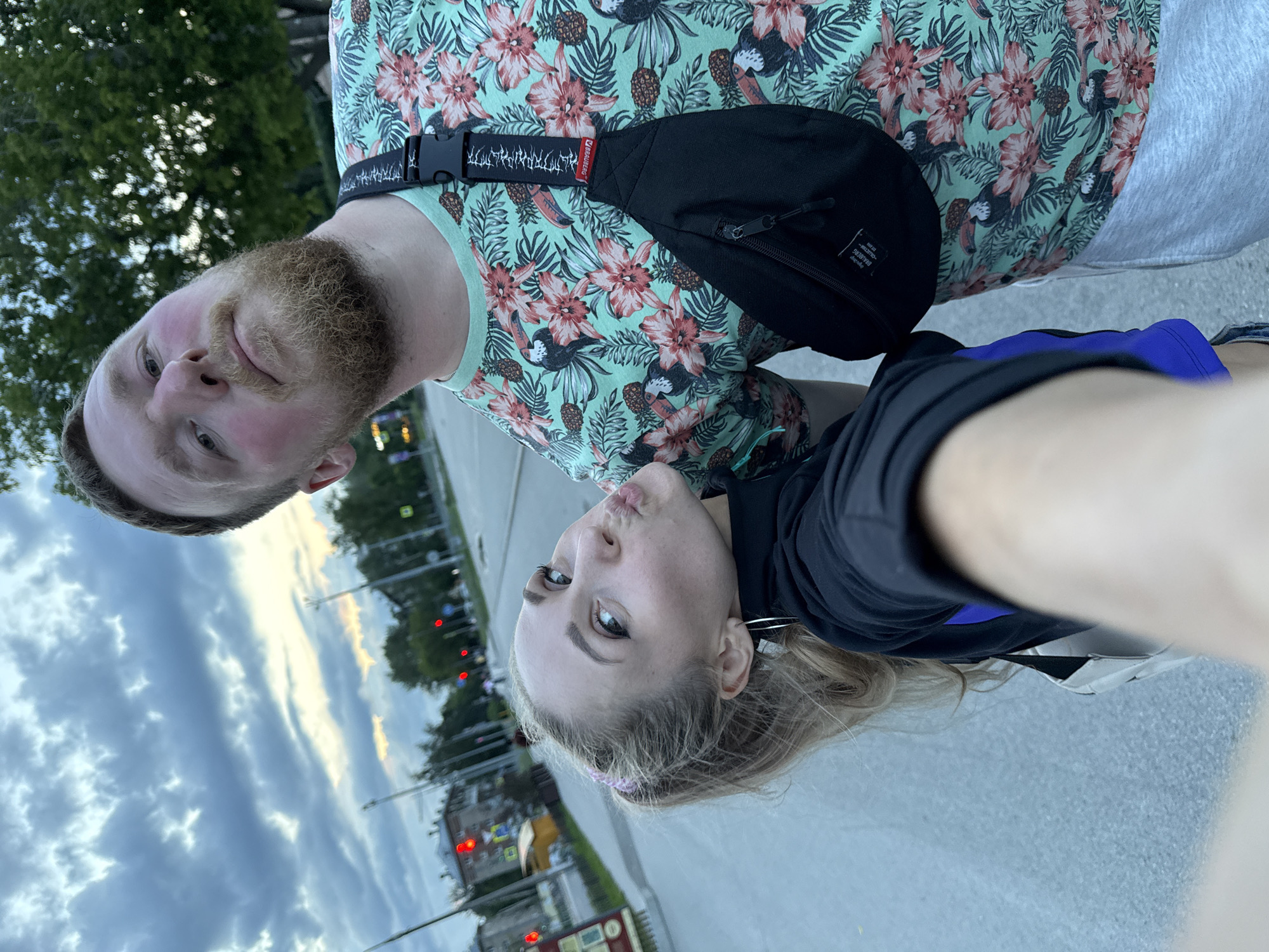 An outdoor, eye-level, close-up, selfie-style shot captures two individuals standing on a paved surface, possibly a sidewalk or road, under a cloudy sky. The scene is set in the city of Perm, Russia, indicated by the presence of distinct, red-lit traffic signals visible in the background, a characteristic of Russian urban traffic control.
On the right, a man with a light-colored beard and fair skin is partially visible, his head tilted back and eyes looking upward. He is wearing a light green t-shirt adorned with a tropical print featuring toucans and flowers, and a black fanny pack is slung across his chest. His cheeks appear flushed.
To the left, a woman with blonde hair pulled back in a ponytail is leaning in towards the camera, puckering her lips. She is wearing a black hooded jacket with a blue lining visible. Her left hand is out of frame, presumably holding the phone to take the picture. Her eyes are directed at the camera.
In the background, trees line the street, and buildings can be seen with windows and red roofs. The road surface is gray and appears wet or reflective. The overall lighting suggests it is either early morning or late evening.