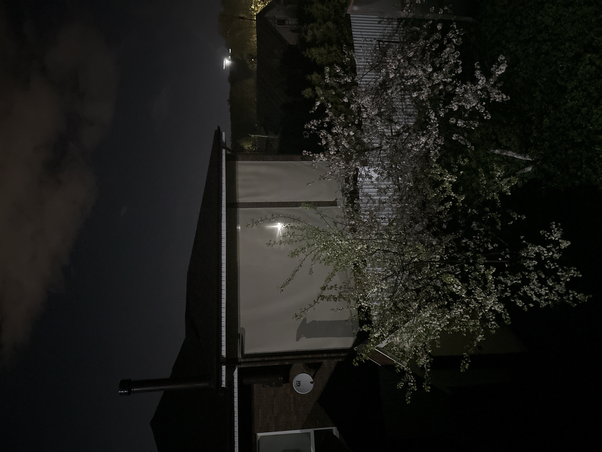 A nighttime scene shows a building exterior. A white wall is illuminated by a light. A tree with white blossoms is visible. A satellite dish is mounted on a brick wall. A chimney is visible. Dark clouds are in the sky. The location is potentially Nizhny Novgorod, Russia, but this is not verifiable from the image itself.