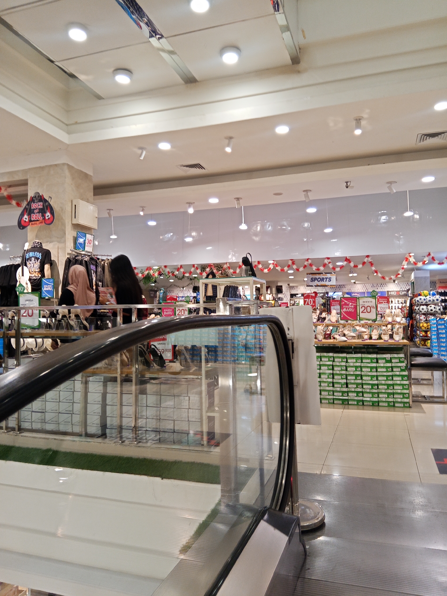 The image captures the interior of a department store, likely in Lubuklinggau, Indonesia, based on the given context. An escalator is prominently featured in the foreground, its metallic handrail curving upwards. Beyond the escalator, the store's layout reveals clothing racks displaying various apparel, including t-shirts and jackets. A prominent "Rock and Roll Music Day" banner hangs on a pillar. Further into the store, a section dedicated to shoes is visible, with displays of footwear and numerous shoe boxes stacked on shelves. A "Sports" sign indicates another departmental section. The ceiling is illuminated with bright, recessed lighting, and decorative garlands of red and white are strung across the store. Two women are present; one, wearing a hijab, is looking at clothing, while the other, with long dark hair, is also in the vicinity of the clothing displays. The overall environment suggests a bustling retail space, likely a department store catering to a range of fashion and sporting goods.