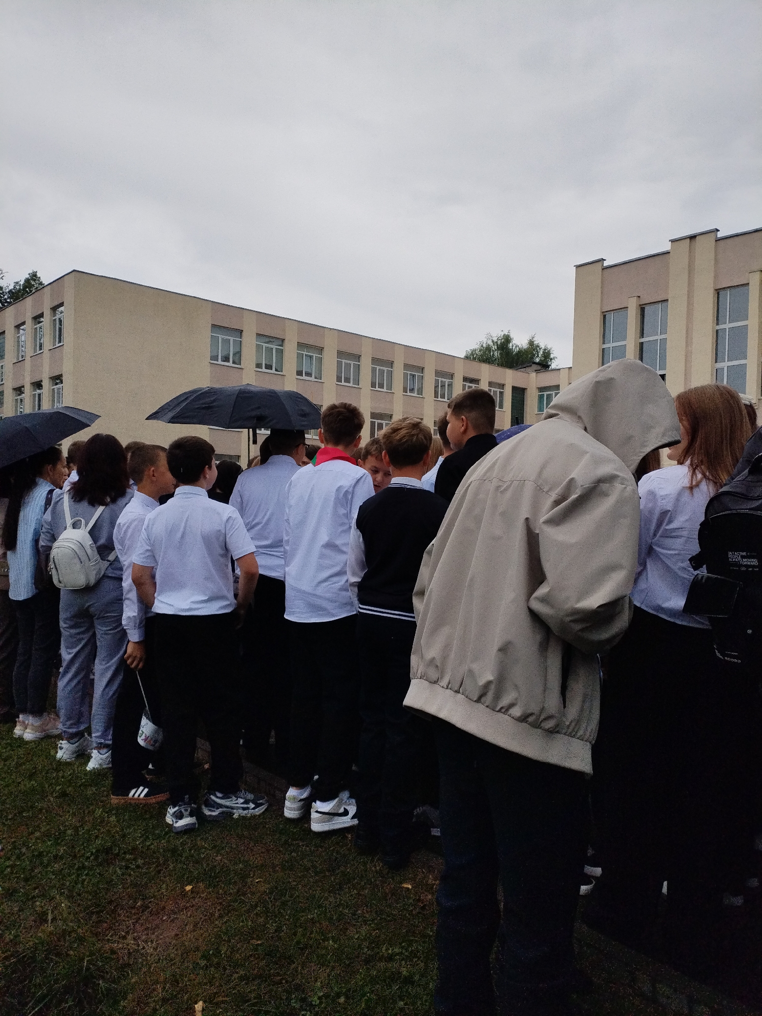 The image depicts a group of young people, likely students, standing in a line outdoors in front of a school building. The sky is overcast and it appears to be raining, as several individuals are holding umbrellas. The school building has a light beige facade with a series of rectangular windows. The students are dressed in what appears to be school uniforms, with many wearing white shirts and dark trousers or skirts. Some are holding backpacks. The ground is covered with green grass and some fallen leaves. The overall impression is a scene of students gathering outside their school on a cloudy, possibly rainy day. The question of the location in Bialynichy, Belarus, cannot be answered from the visual information provided in the image.