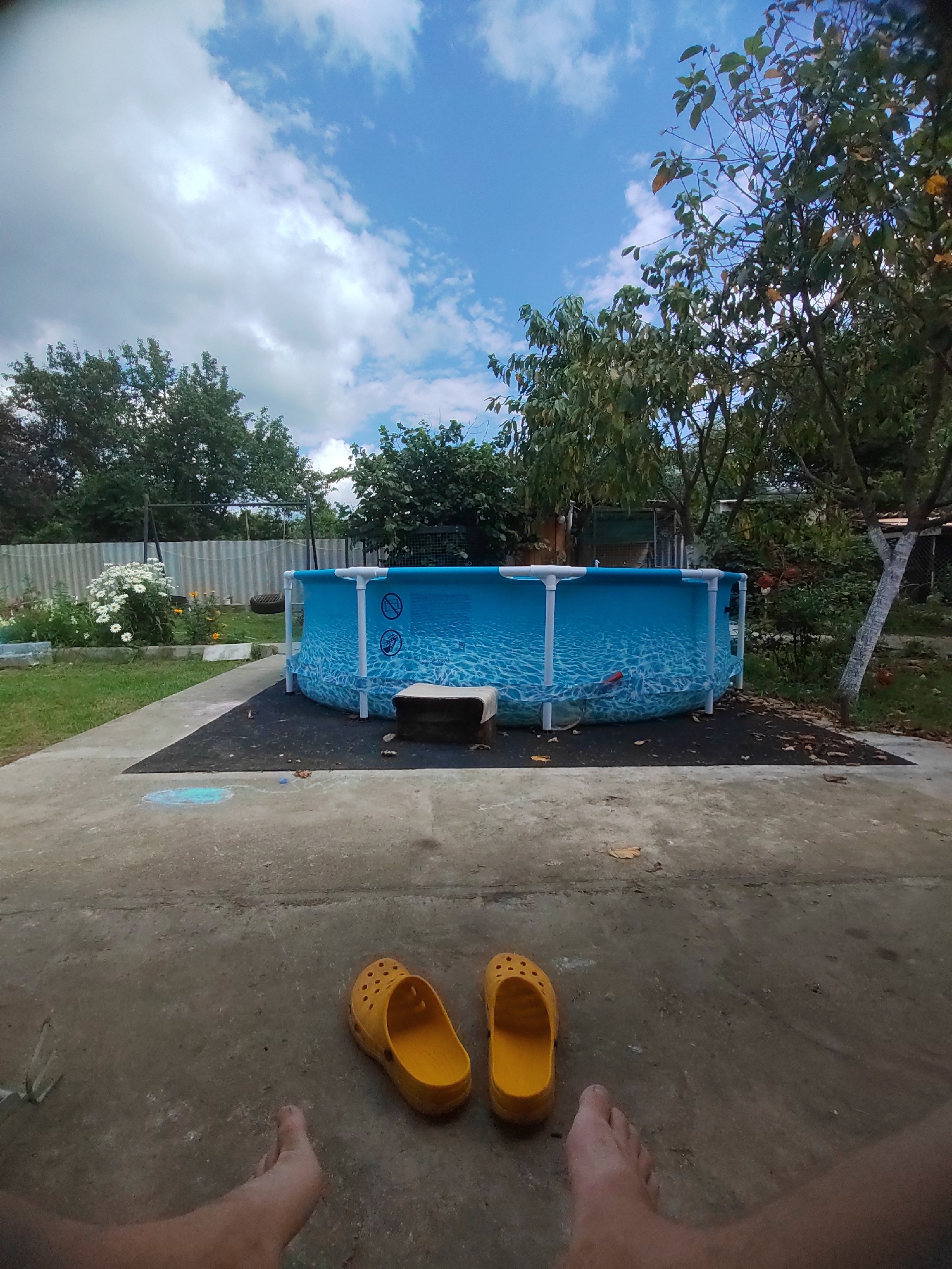 The image captures a backyard scene on a cloudy day in Psebai, Russia. In the center of the frame, an inflatable blue swimming pool with a white frame stands on a dark rectangular mat. The pool is filled with water, and its sides display a pattern of waves. To the left of the pool, a garden bed with white flowers and green foliage is visible, bordered by a corrugated metal fence. A swing set with a tire swing is also present in the background. The foreground shows a concrete patio where a pair of yellow clogs and two bare feet are positioned. Trees with green and some yellowing leaves surround the yard, and a white tree trunk is prominent on the right side of the image. The sky is a mix of blue and white clouds.