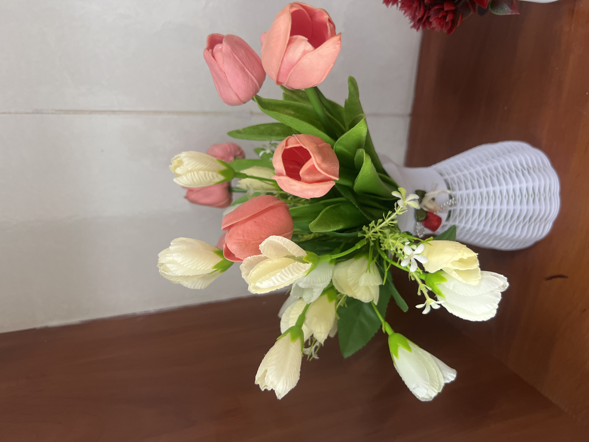 A close-up, eye-level, indoor shot features a white woven vase filled with artificial tulips. The bouquet is comprised of several pink and cream-colored tulips, interspersed with green leaves and small white flowers. The vase sits on a dark brown wooden surface, and behind it, a wall with a wood-paneled section and a tiled section is visible. The image does not contain any information that would allow for its location in Nookat, Kyrgyzstan.