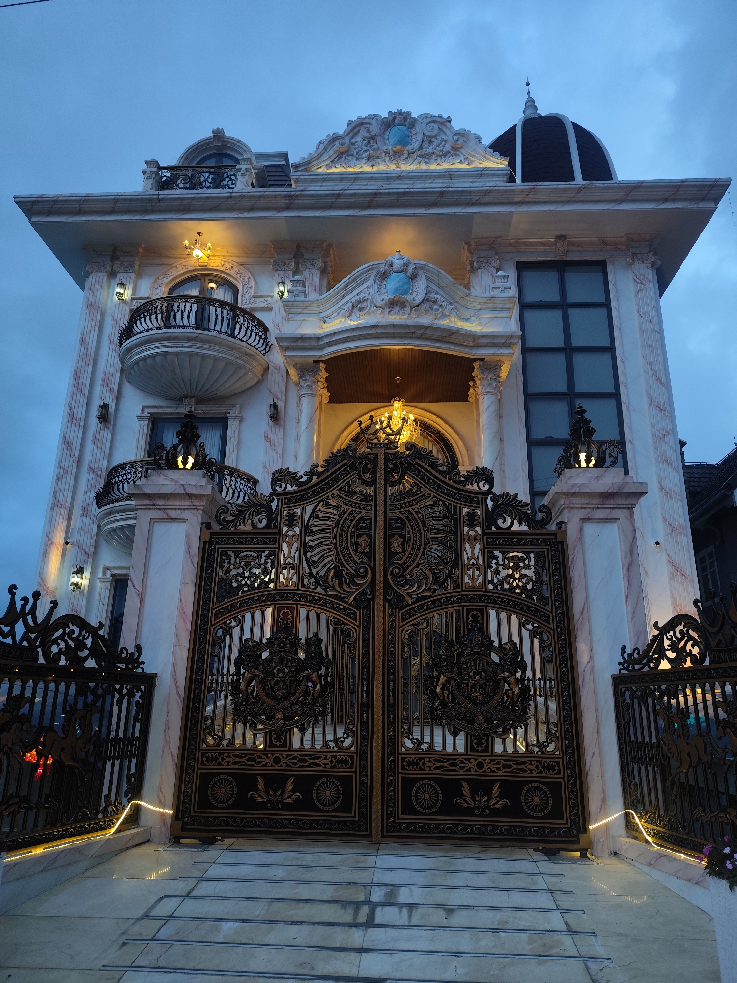 The image shows an imposing, multi-story mansion with ornate architectural details, located in the city of Xuân Trường - Đà Lạt, Vietnam. The mansion features a dark dome on its roof, classical-style balconies with wrought-iron railings, and decorative moldings. The facade is primarily white with marble-like pink veining. A large, elaborate black and gold gate with intricate scrollwork and a crest guards the entrance. The gate is illuminated by yellow string lights along the bottom and a chandelier is visible within the entryway. The exterior is further accented by decorative lamps and what appears to be a security camera on the right-hand pillar. The sky is overcast and appears to be twilight. There are no people visible in the image. The foreground shows a set of wide, stone steps leading up to the gate.