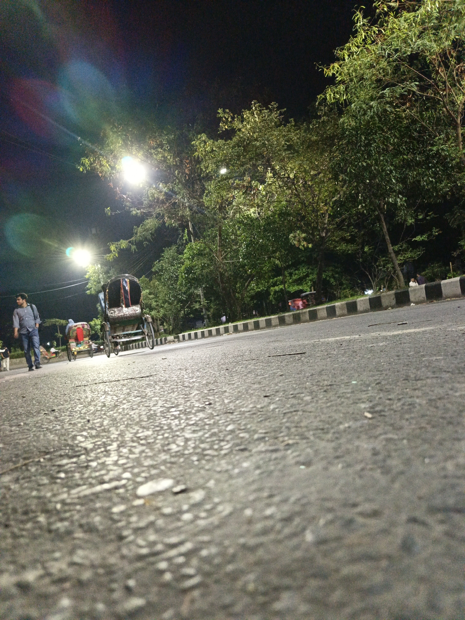 The image depicts a street scene at night, likely in Dhaka, Bangladesh, based on the presence of cycle rickshaws. The focus is on the asphalt road, which takes up most of the frame with a low-angle perspective. To the left, a man walks along the road, wearing a light-colored shirt, dark pants, and carrying a backpack. Two cycle rickshaws are visible: one directly in front, in focus, and another slightly further behind and to the left. Streetlights illuminate the scene, creating glare in the upper left corner. Along the right side of the road is a curb with alternating black and gray blocks, and beyond that, trees line the street. In the distance, a red vehicle can be seen, along with indistinct figures of people. The overall impression is that of a street scene at night with pedestrians and cycle rickshaws.