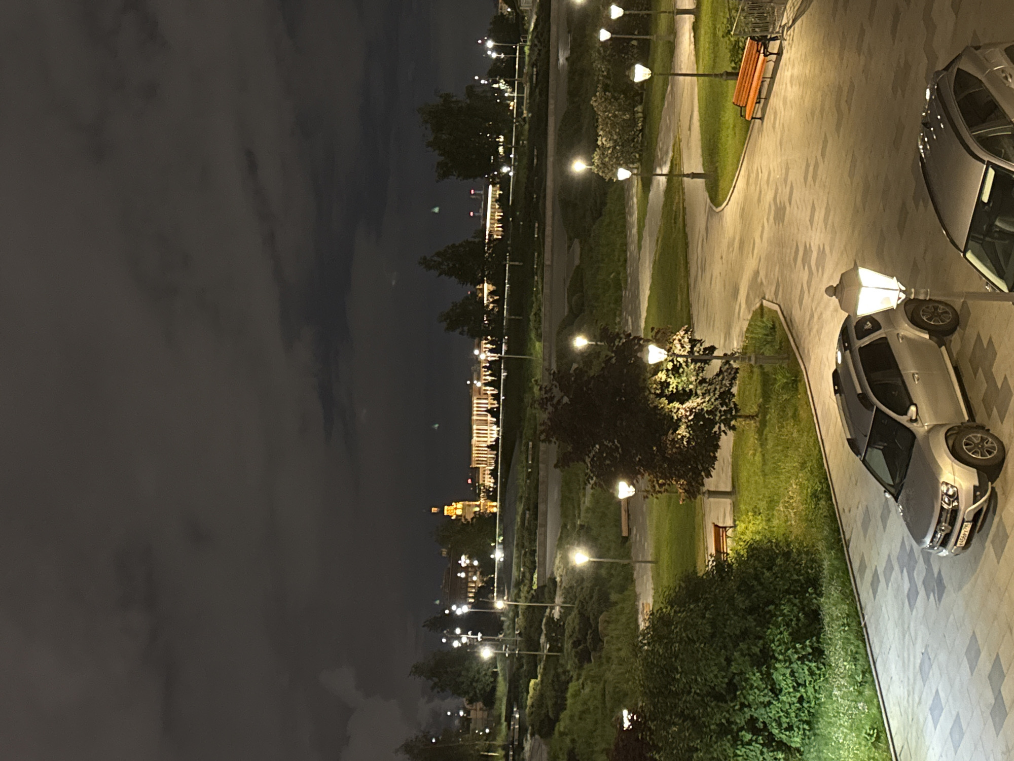 The image depicts a nighttime scene in Moscow, Russia, likely a park or landscaped area along the Moskva River. Buildings are visible in the distance, illuminated. A pathway with benches and lampposts is present, with two vehicles parked on the right. One is a silver SUV, the other a dark colored car. The sky is overcast. No people are directly visible.