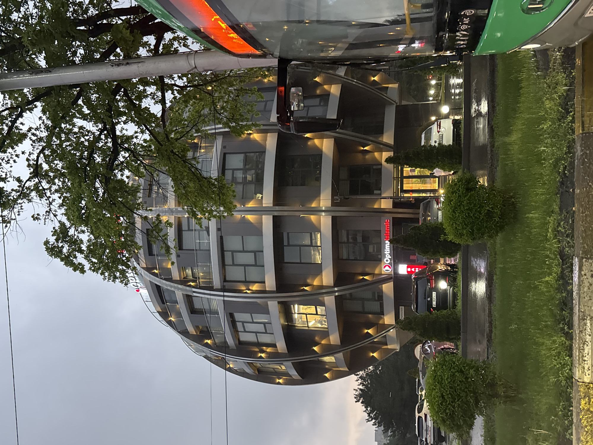 This image captures an urban street scene in Bishkek City, Kyrgyzstan, likely during the evening or late afternoon after a rainfall, given the wet pavement and reflections.
The central subject is a modern, multi-story building with a distinctive curved, almost oval facade. The building's exterior is dark grey or black, featuring horizontal bands that incorporate upward-facing lights, illuminating sections of the structure. Numerous large windows are visible, with some interior lights on. A large tree with green foliage partially obscures the building on the left.
In the foreground and to the right, a wet road surface reflects ambient lights. Several cars are parked along the road, including a dark SUV with its taillights illuminated and a white car further down. A large green and black bus is partially visible at the top, suggesting it is either passing by or parked nearby. A well-maintained grassy median with small, trimmed bushes separates the road from a sidewalk area.
Visible text on a prominent red vertical sign on the building reads "OptimaIslamic". A vehicle license plate is partially visible, showing "009..." and another with "909J...". The sky above is overcast, contributing to a muted, calm atmosphere. The scene depicts a typical urban environment during a transitional light period, with a focus on modern architecture and infrastructure.