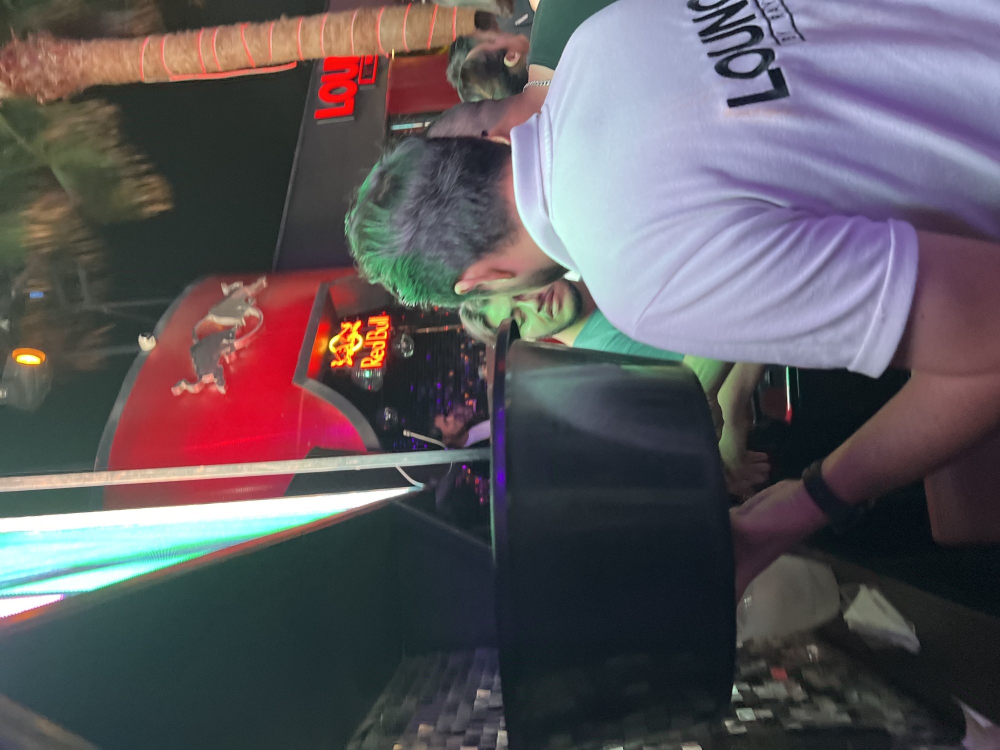 The image shows a crowded, outdoor scene at night, with signage indicating "Lounge" and "Red Bull." A partially visible palm tree and a building facade are illuminated with red and blue lights. Several individuals are present. One person, wearing a white t-shirt with black writing is in the foreground, partially obscuring the view. A person in a green shirt is visible behind them. The location is Arica, Chile.