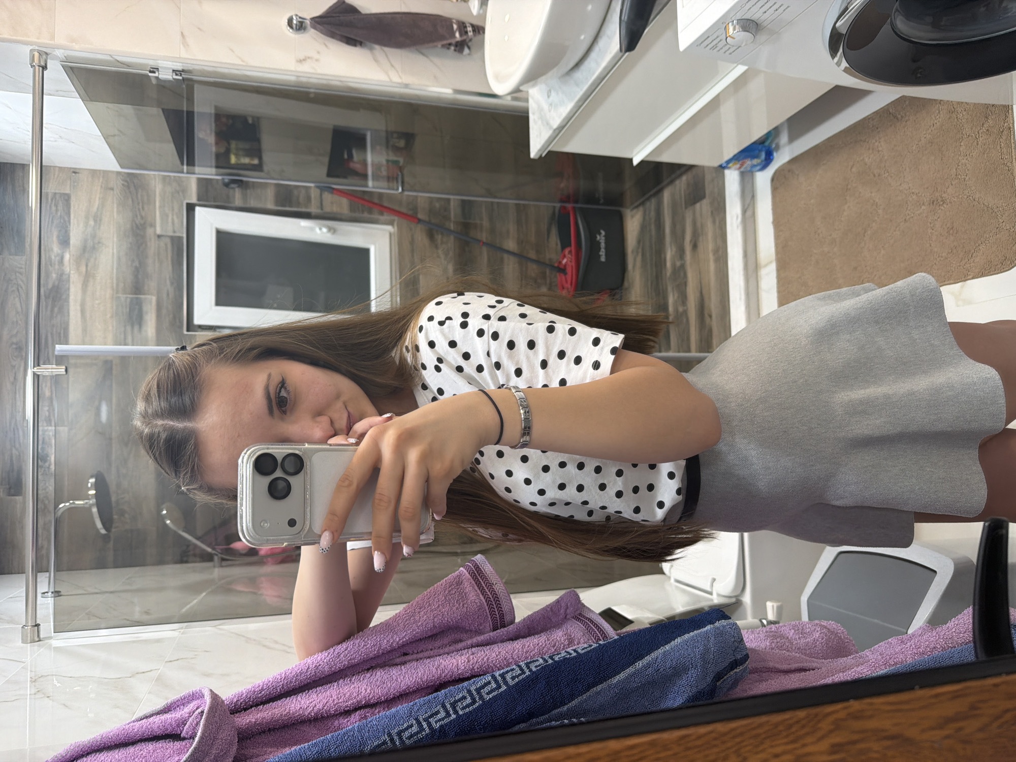 A young woman is taking a selfie in a bathroom. She is wearing a white shirt with black polka dots and a gray skirt. Her hair is long and brown. She is holding a silver iPhone with a clear case. The bathroom has a shower with a glass door, a white sink, a washing machine, and a brown rug. Towels are draped over the edge of what appears to be a counter or shelf. The background appears to show elements of a home environment. The location is specified as Buhovo, Bulgaria.