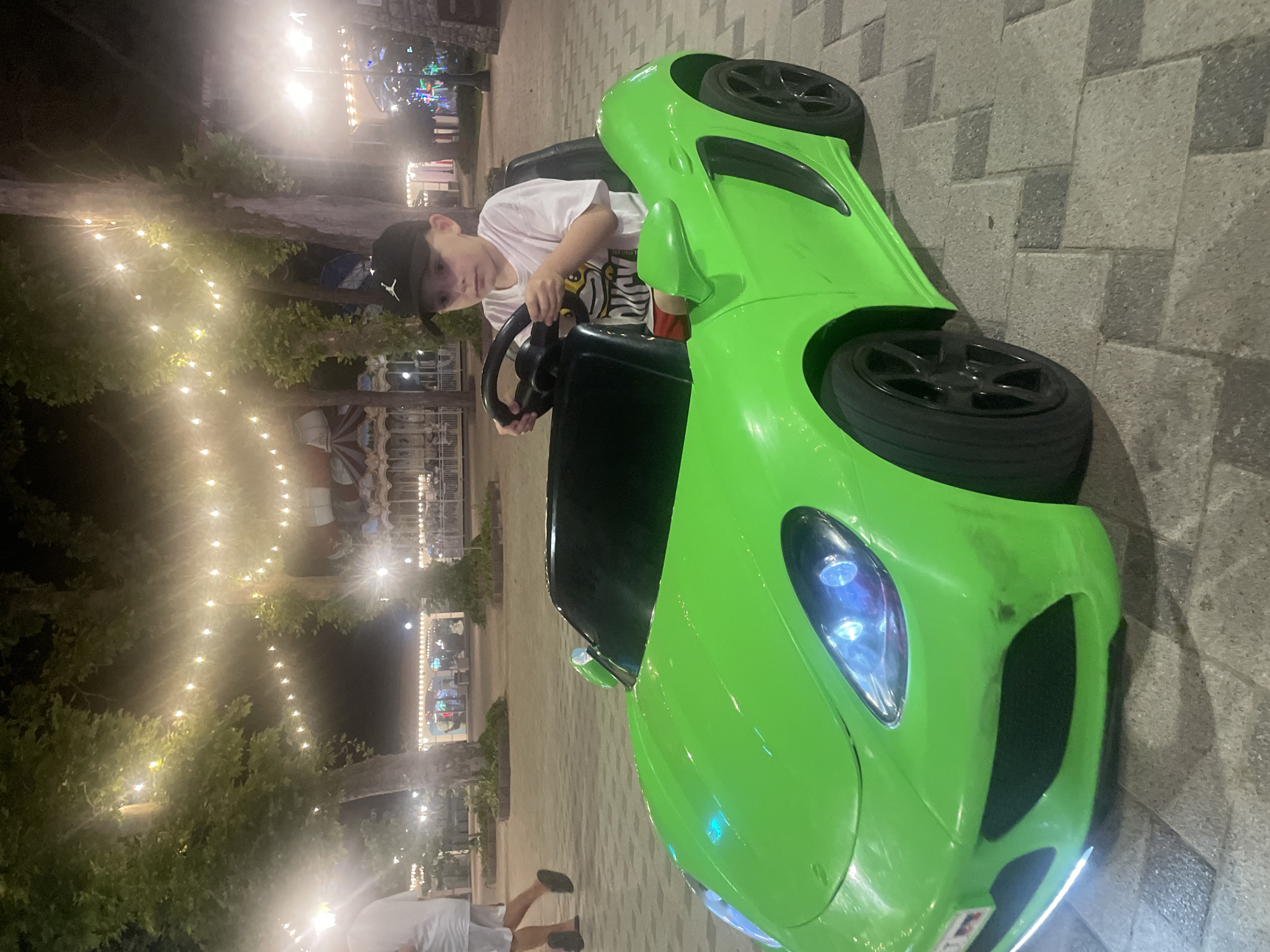 A young boy wearing a white t-shirt and a black baseball cap is sitting in a bright green toy car. He is holding the steering wheel. The car is on a paved area with square stones. In the background, there are trees with string lights overhead. A carousel and other amusement park rides are visible in the distance. A person in a white shirt and shorts is walking away from the camera on the left side of the image. The scene appears to be at an amusement park or a similar entertainment venue in the city of Anapa, Russia, likely at night due to the artificial lighting.