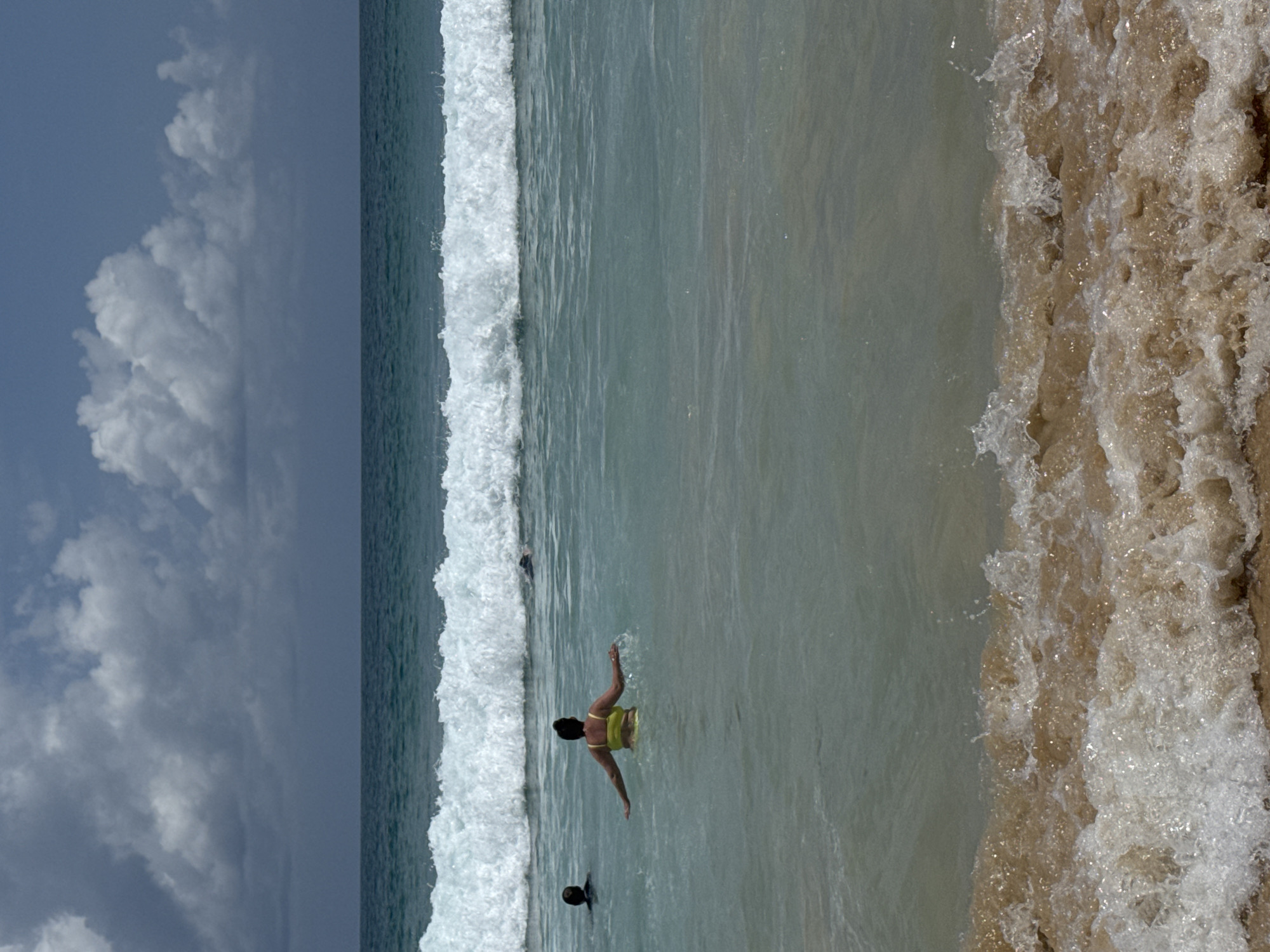 The image shows a view of the ocean in Rawai, Thailand, with a person swimming in the water. The sky is partly cloudy, with a mix of dark and white clouds. The ocean is a clear, turquoise color, with a large wave breaking in the background. In the foreground, a person in a yellow bikini is standing in the water, with their arms outstretched. Two other people are also visible in the water, further out. The beach is a light brown color, with some white foam from the waves.