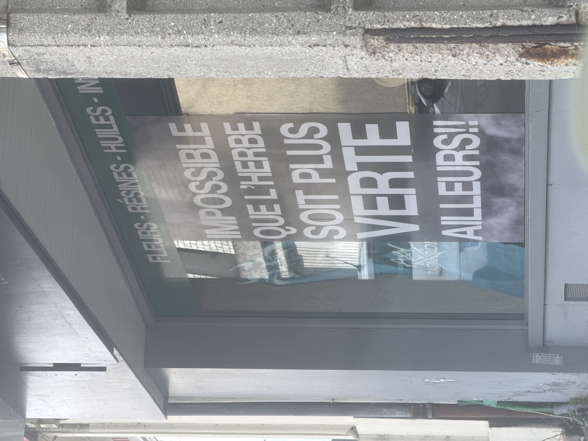 The image, rotated 90 degrees clockwise, shows a storefront window and part of a building facade in what appears to be an urban setting, consistent with Orsay, France.
Above the large display window, a green sign with white capital letters reads: "FLEURS - RÉSINES - HUILES - IN". The "IN" is incomplete, likely indicating "INFUSIONS" or a similar product category.
Inside the window, a prominent poster displays bold white text on a dark grey background. The text reads: "IMPOSSIBLE QUE L'HERBE SOIT PLUS VERTE AILLEURS!!", which translates to "Impossible for the grass to be greener elsewhere!!". This is a marketing slogan suggesting the superior quality of the establishment's offerings. A smaller, partially visible logo or brand name, "RAGYARON," is also present on the poster. Reflections on the window obscure some details of the interior.
The building's exterior primarily features grey concrete, with some textured sections and visible rebar near the top. In the bottom right corner, a small white sticker on the concrete displays a phone number, "09 72 10 34 48," with other faded, unreadable text. Below the window, pipes or cables are visible. The scene appears to be set during the daytime, with no individuals or specific activities actively depicted.