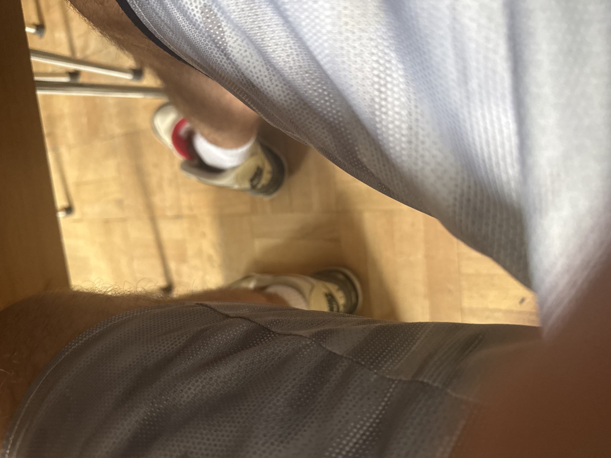 This is a close-up, low-angle shot of a person's legs wearing grey athletic shorts and white socks with beige and red athletic shoes. The person is seated at a wooden table, as indicated by the underside of the table in the upper part of the frame and its legs in the background. The floor appears to be light-colored wood paneling. The image provides no specific details about the location within Sofia, Bulgaria, beyond the presence of indoor furniture and flooring.