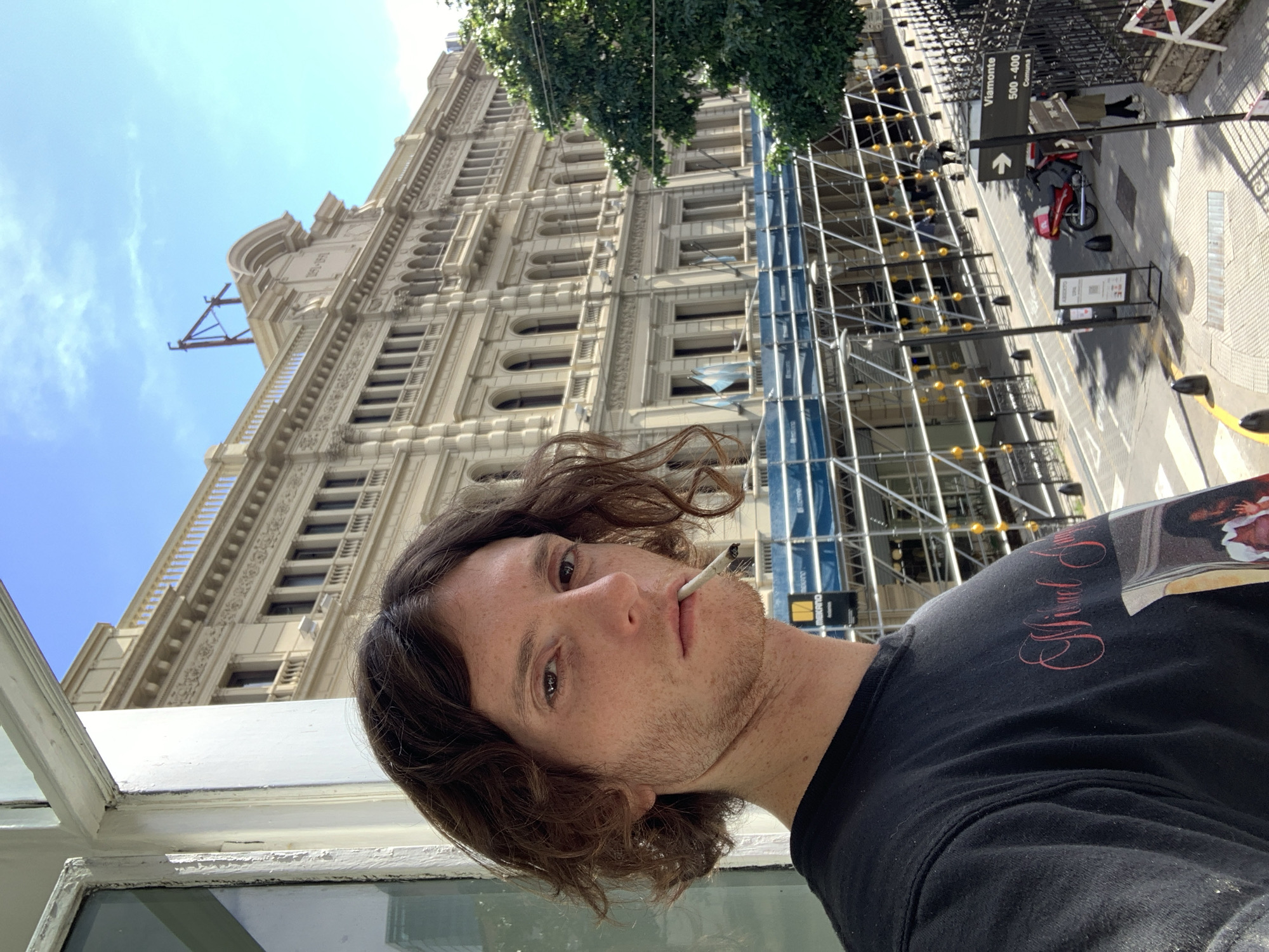 A low-angle, medium shot depicts a man with dark hair and stubble, looking directly at the camera with a cigarette in his mouth. He is wearing a black t-shirt with a graphic print on the shoulder. Behind him, an ornate, beige-colored building rises several stories high. Scaffolding is erected against the building's façade. A street sign indicating "Viamonte 500-400 Comuna 1" is visible on the right side of the frame, indicating the location is in Buenos Aires, Argentina. A sidewalk and a parked red motorcycle can be seen in the background. The sky is blue with scattered clouds. The man appears to be standing on a balcony or near a window.