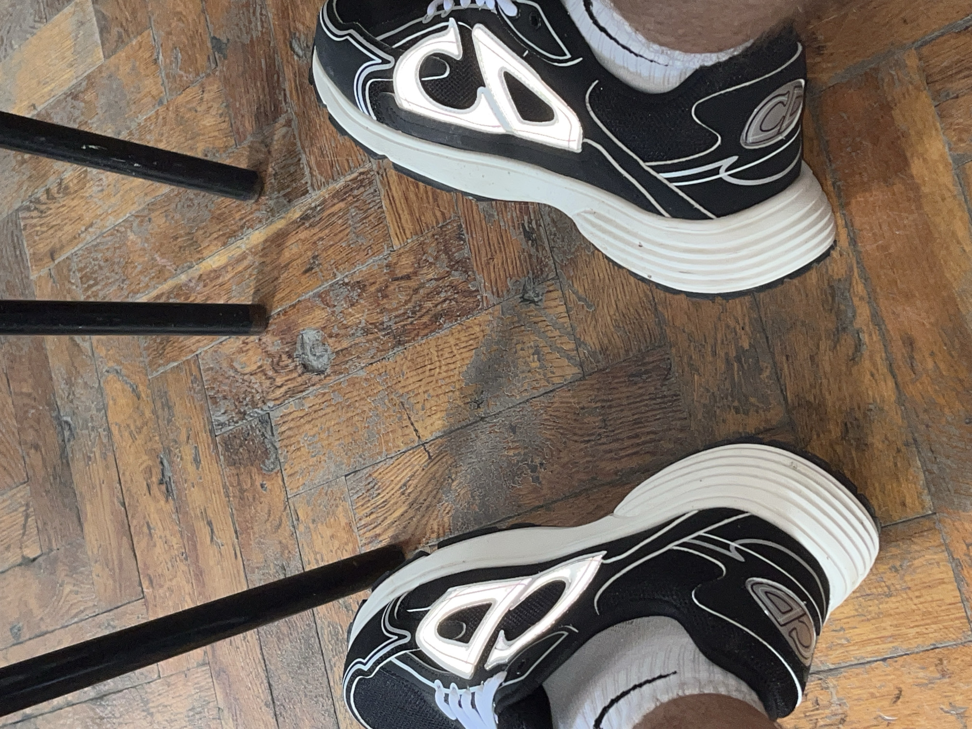 The image shows a pair of black and white athletic shoes on a wooden floor. The shoes have reflective accents and white soles. Two dark metal chair legs are visible in the background. The scene appears to be indoors, possibly in a residential setting. There is no indication of the location being in Zlatograd, Bulgaria, or any other specific city.