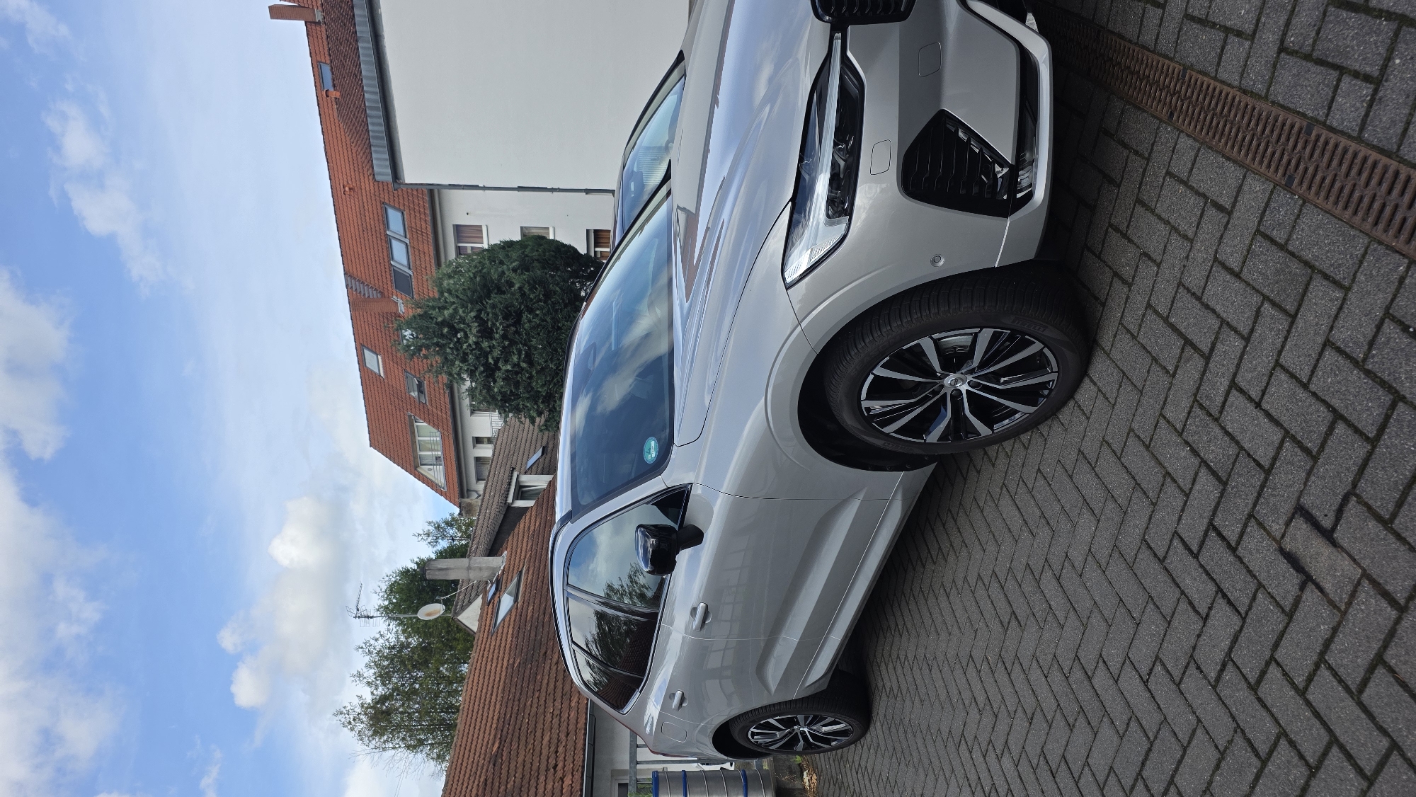 A silver Volvo XC60 is parked on a herringbone pattern brick driveway. The SUV is positioned at an angle, facing slightly away from the camera. The car features black and silver multi-spoke wheels. In the background, there are red-tiled roofs of residential buildings and some green trees. The sky above is partly cloudy with a mix of blue sky and white clouds. The scene appears to be in a residential area.