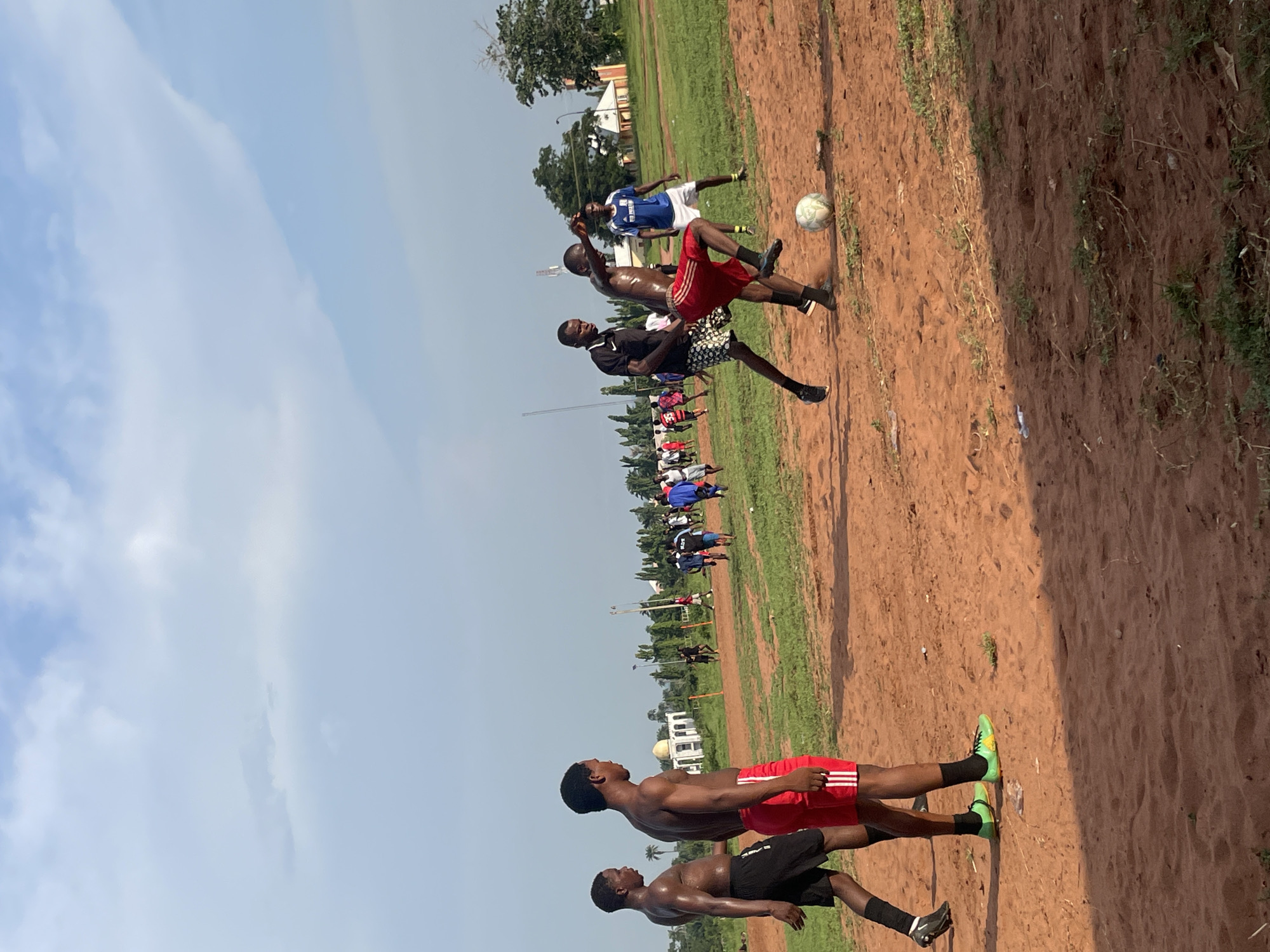 The image captures an active outdoor scene on a sunny day in Ogbogbo, Nigeria, depicting a casual soccer match or practice. The main playing area is an unpaved reddish-brown dirt field with scattered patches of green grass. A clear blue sky with some white clouds dominates the upper portion of the frame.
In the mid-ground, a white soccer ball is in the air, surrounded by three players actively engaged in play. One player, shirtless and wearing red shorts, is mid-jump, seemingly having just kicked or heading towards the ball. Another player, in black shorts and a black top, is positioned with his foot near the ball. Behind them, a player in a blue jersey and white shorts is also focused on the action.
In the foreground, to the left, two shirtless young men stand, observing the game. One is muscular, wearing red shorts with white stripes and bright green soccer cleats. The other, also muscular, wears black shorts and black cleats. Both appear focused on the field.
In the background, beyond the immediate play, numerous other individuals are visible on the field, some wearing jerseys, suggesting a larger gathering or multiple teams. Further behind them, lush green trees and several buildings, including one with a distinctive dome, line the horizon. Goalposts are also faintly visible, confirming the setting as a soccer pitch.
The time of day appears to be bright daylight, possibly morning or late afternoon, given the clear sky and illumination. The players exhibit focus and exertion. There is no visible text discernible in the image.