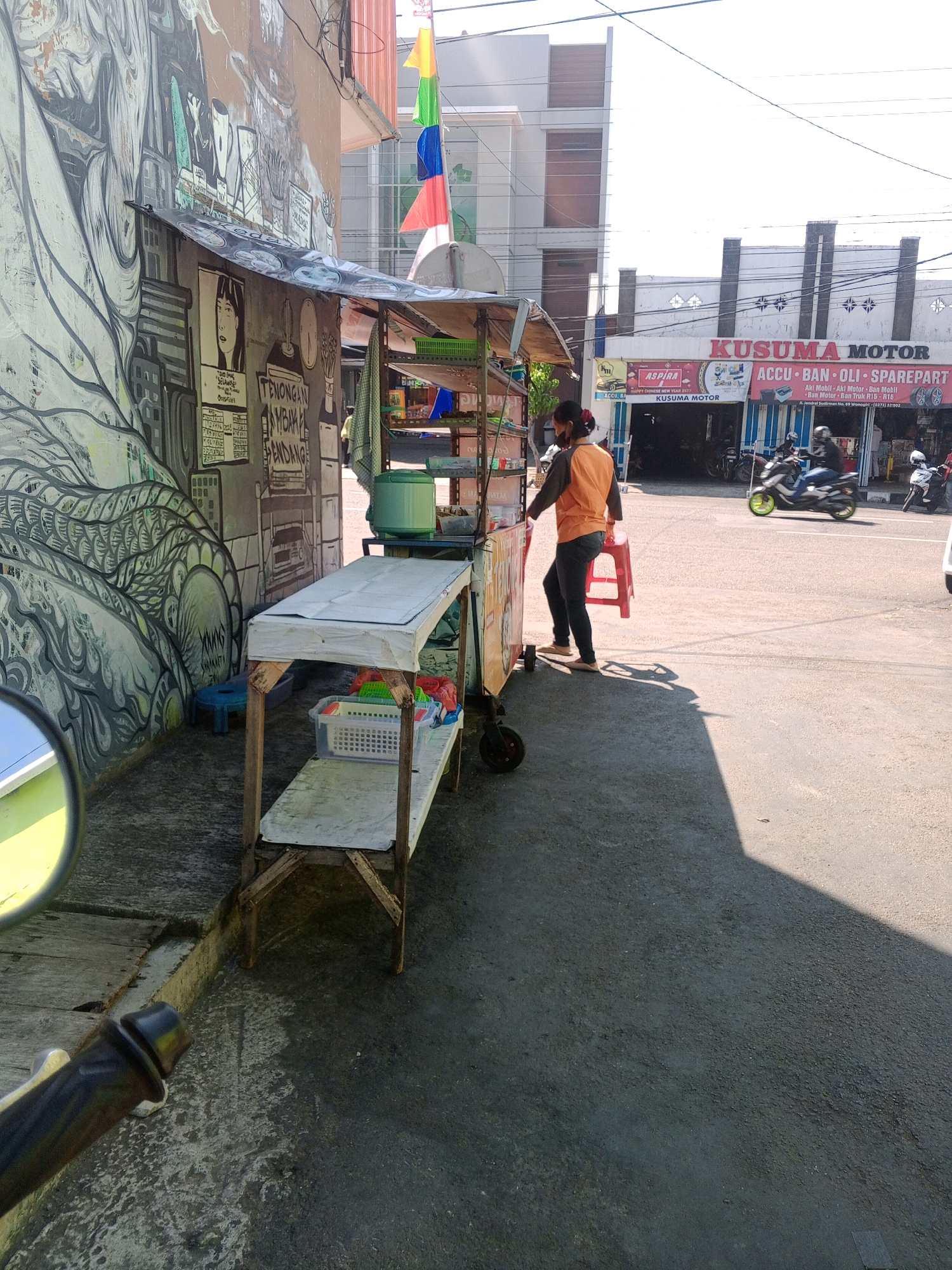 An outdoor, daytime scene in Wonogiri, Indonesia, features a small roadside stall with a person operating it. The stall is adorned with a colorful banner featuring flags of red, yellow, green, and blue. To the left of the stall is a wall covered in graffiti. A person wearing an orange shirt and dark pants stands near the stall, holding a red plastic stool. In the background, a building with the sign "KUSUMA MOTOR" is visible, indicating a motorcycle repair shop. Several motorcycles are parked in front of the shop, and a few people are present. The ground is paved, and sunlight casts shadows. The perspective of the image is from a low angle, possibly from a motorcycle.