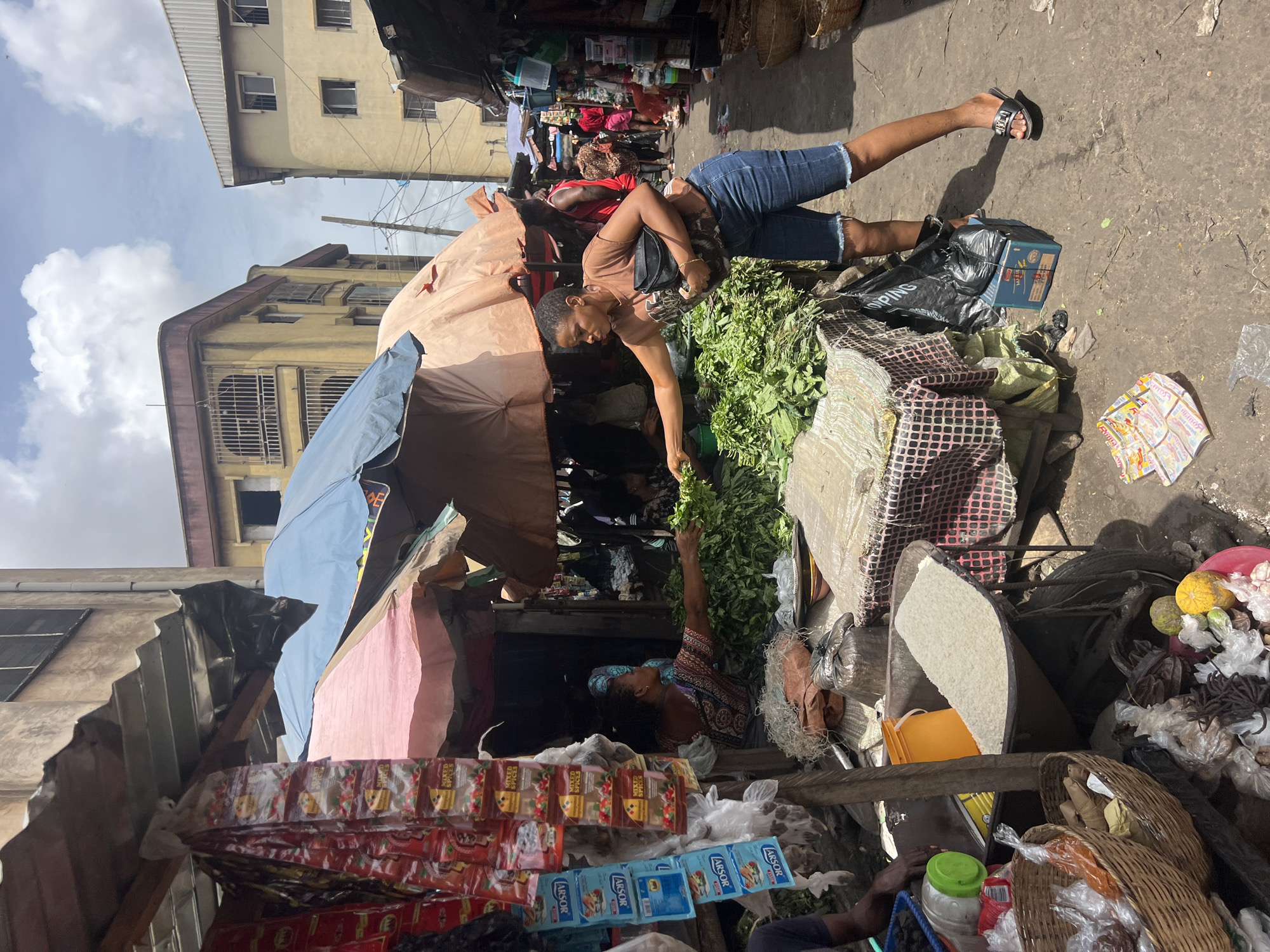 The image depicts a bustling marketplace in Nkpor, Nigeria. A woman wearing a brown shirt and denim shorts stands near a stall, reaching for a handful of leafy greens. She has a black bag over her shoulder and is wearing sandals. Another woman, wearing a patterned blouse and headscarf, is behind the stall and also reaching for the greens. The stall is covered with various tarps and fabrics. A variety of goods, including prepackaged spices and other food items, are displayed at the front. Several baskets, a bag of white rice, and various fruits and vegetables are arranged nearby. The ground appears to be a paved surface with some debris. Buildings are visible in the background. Other people are present in the market, moving through the scene.