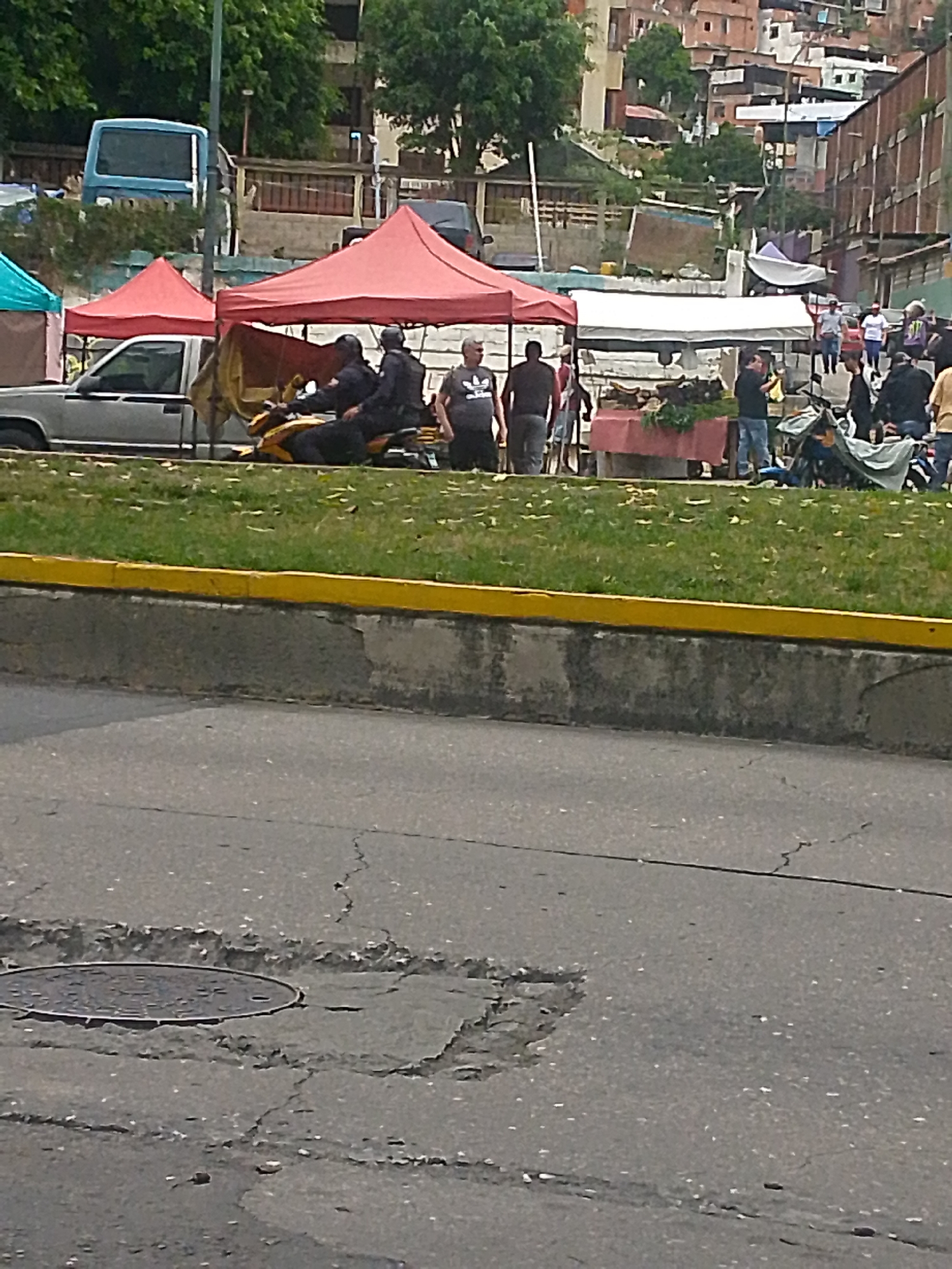 The image shows a street scene in Caracas, Venezuela. The foreground features a cracked asphalt road with a manhole cover. Behind the road is a raised grassy area with a yellow-painted curb. Along the back of the grassy area are several tents with red and white tops, suggesting a market or street fair. People are milling around the tents, some standing at tables, while others walk by. Two people in what appears to be police uniform are sitting on a yellow motorcycle parked near the tents. A gray pickup truck is parked nearby. The background features densely packed buildings on a hillside, typical of Caracas. The overall impression is of a busy urban scene with street vendors and activity.