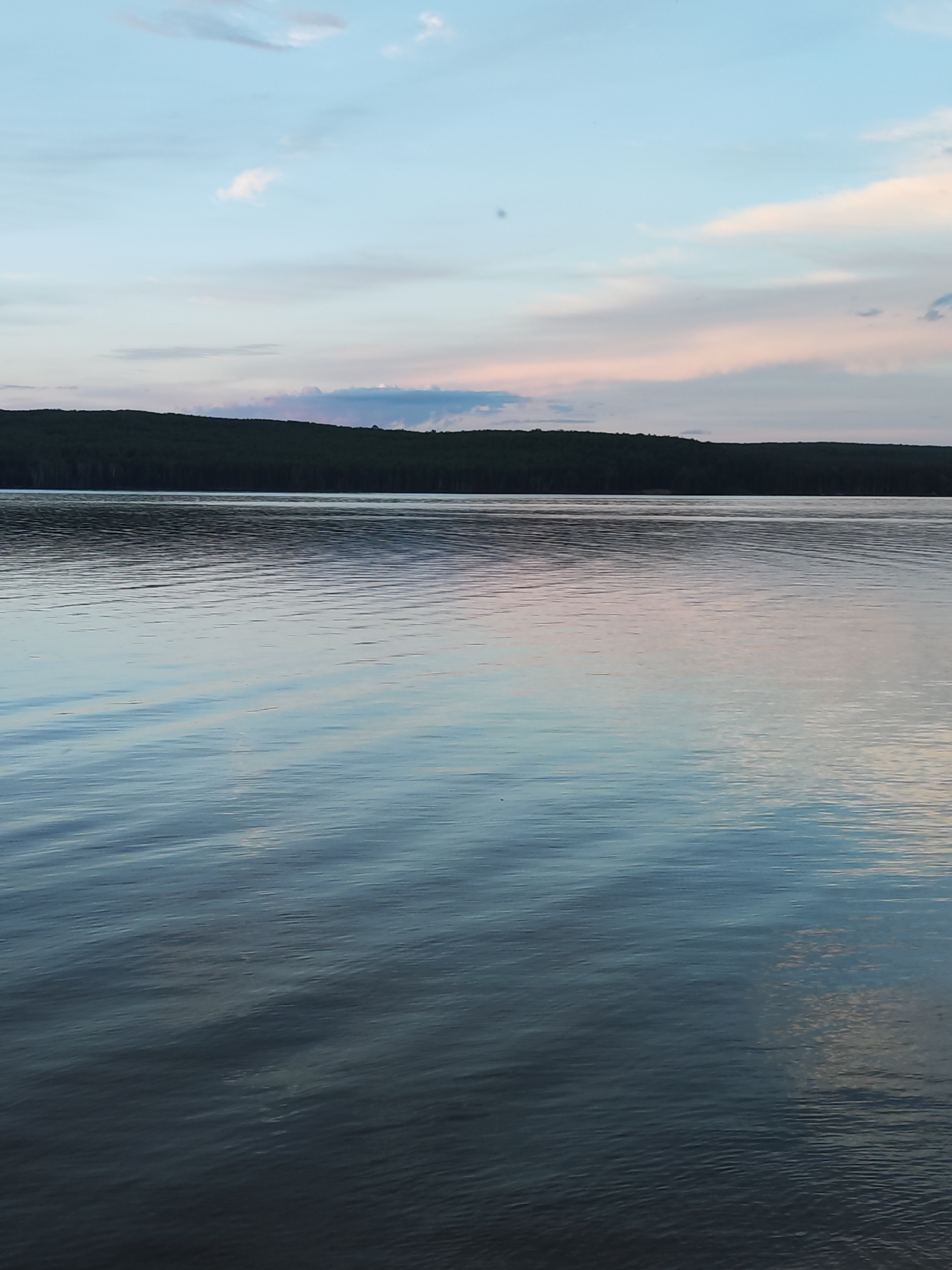 The image depicts a body of water, possibly a lake or river, with a treeline along the horizon under a sky with blue and pink-tinged clouds. The water is rippled, reflecting the colors of the sky. The location is indicated as Chaikovsky, Russia, suggesting the body of water could be part of the Kama River or a nearby reservoir. There are no people, subjects, or actions visible in the image.