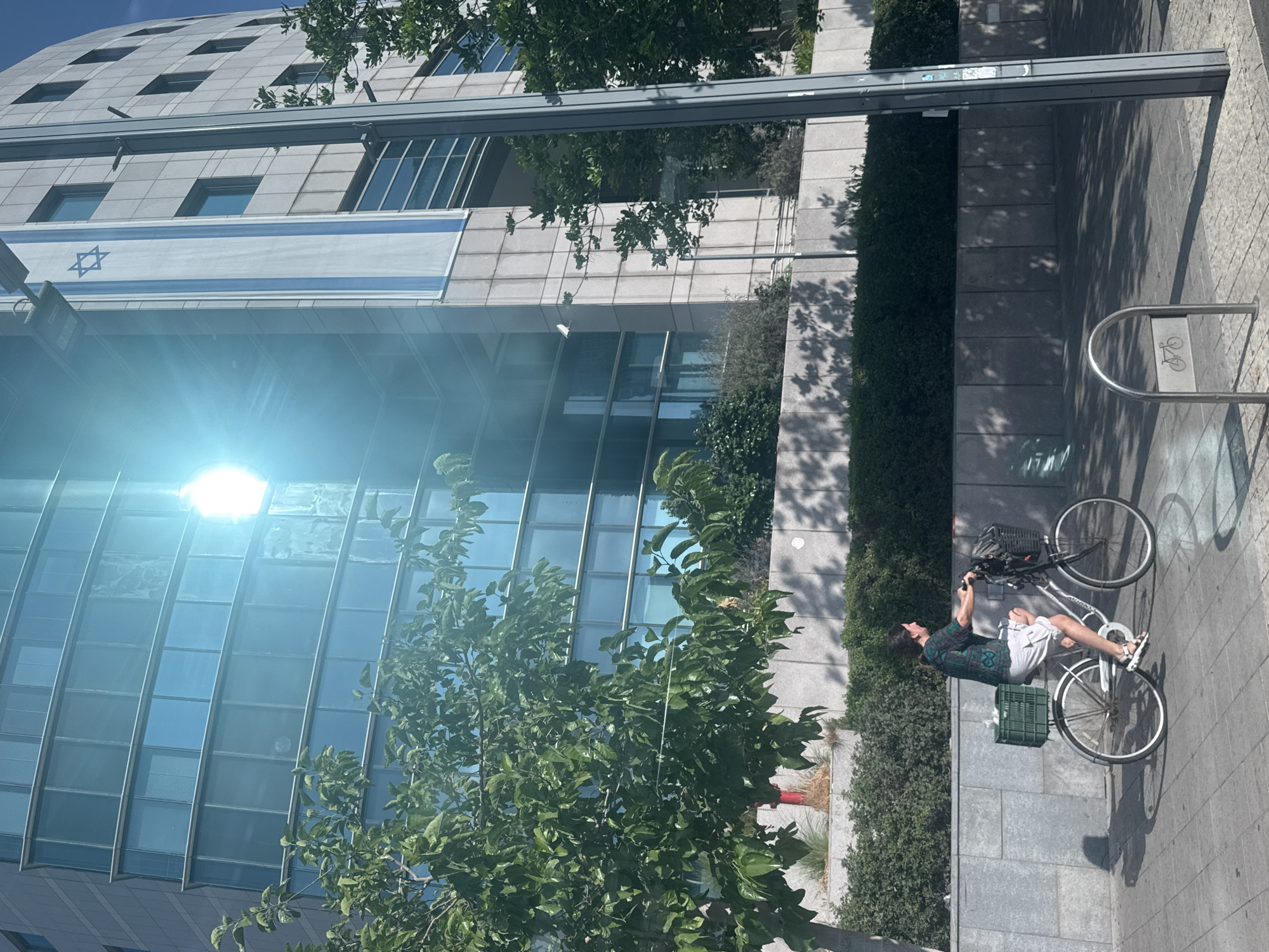 A person is riding a bicycle on a sidewalk in front of a modern building. The building has a facade of glass and stone, and an Israeli flag is hanging on its side. The person on the bicycle is wearing a patterned shirt and light-colored shorts. The bicycle has a green basket on the back and a black basket on the front. The sun is shining brightly, creating a lens flare effect in the image. The background includes trees and bushes. This is likely in Tel Aviv, Israel, given the presence of the Israeli flag.