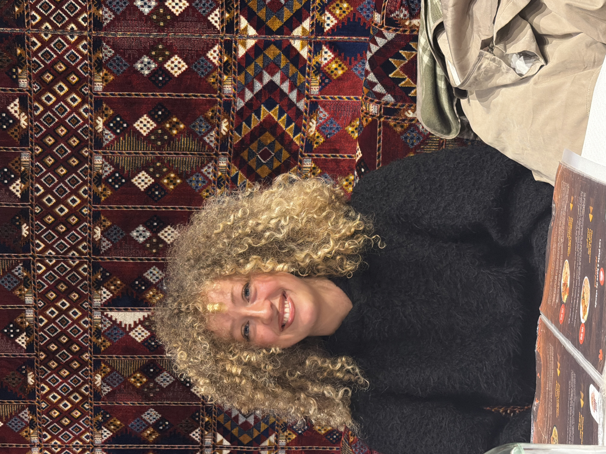 A woman with curly blonde hair is smiling at the camera while seated next to a table in an indoor setting. She is wearing a dark, textured sweater. To her right, a menu is visible, partially obscured. Behind her is a patterned rug with red, gold, and blue geometric designs. In the upper right corner, there is a light-colored jacket or coat draped over something. The location is possibly Paris, France, based on the style of menu and overall aesthetic.