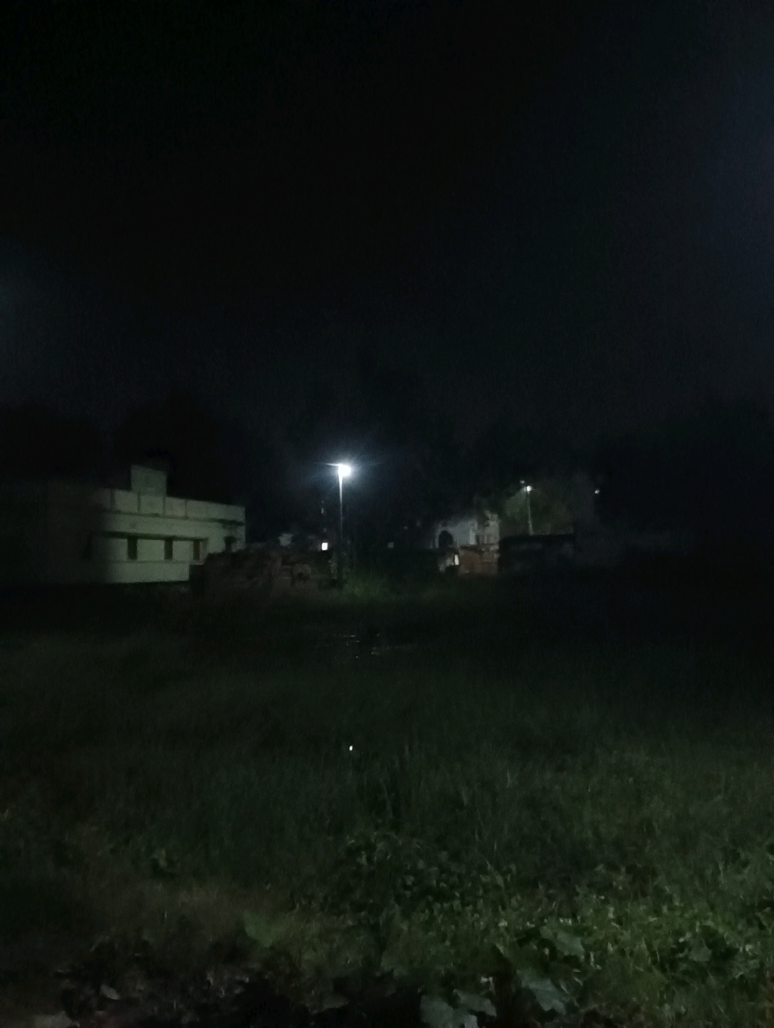 This image depicts a nighttime scene in Dakhin Parsa, India. In the mid-ground, a two-story building with a light-colored facade and rectangular windows is visible on the left. To the right of the building, a tall, slender pole with a lit lamp stands, illuminating the surrounding area. A pile of what appears to be stacked material, possibly hay or wood, is situated near the building and the lamp post. The foreground is dominated by dense, dark green grass, suggesting an unkempt field or undeveloped land. The overall ambiance is dark and somewhat ominous, typical of a rural or semi-urban setting at night, with limited light sources. The sky is completely black, indicating the absence of daylight.