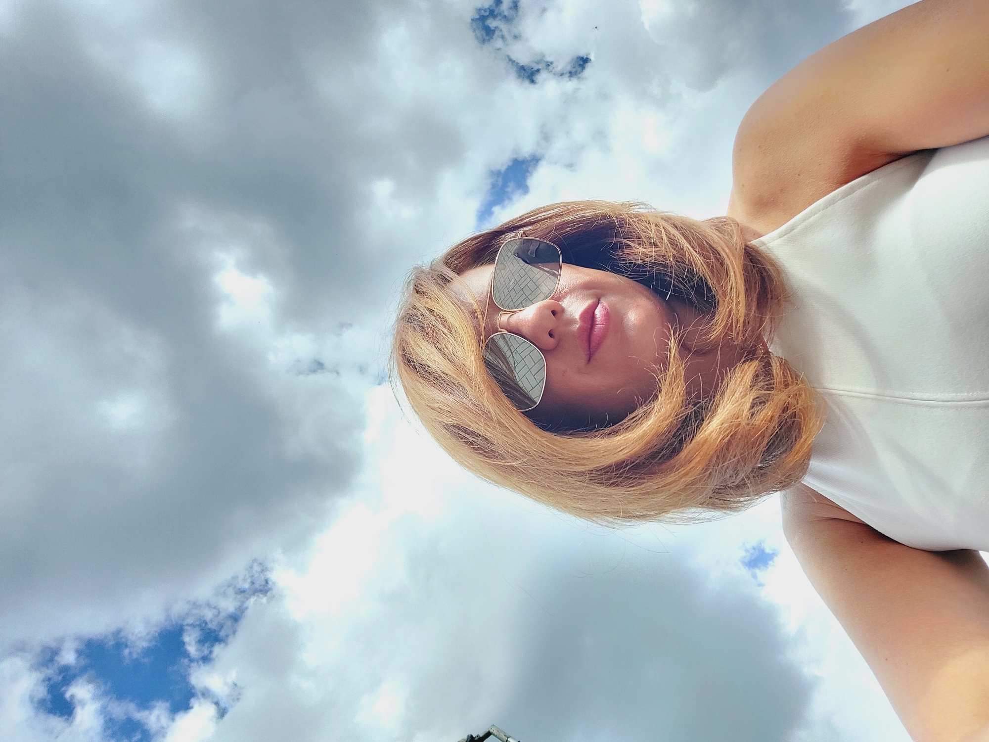 An outdoor, low-angle, eye-level, medium shot shows a woman with auburn hair and sunglasses looking down and to the right against a sky with dramatic clouds. The woman is wearing a white halter top and her face is in focus. The clouds are gray and white, with patches of blue sky visible. The scene is likely in Sofia, Bulgaria, given the context provided, though there are no specific visual cues in the image to confirm this location. The overall lighting suggests it is daytime, possibly overcast.
