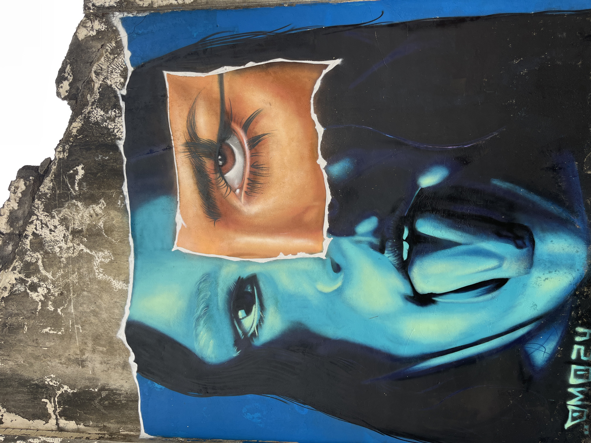 The image shows a piece of street art on a concrete wall. The artwork depicts a stylized face, split into two distinct sections. The left side of the face is rendered in shades of blue and teal, with a portion of an open mouth and a protruding tongue. The right side of the face is shown in warmer tones of orange and brown, focusing on a single eye with dramatic eyeliner and long eyelashes. The artwork is applied to a weathered and cracked concrete surface, with the top edge of the image showing the rough texture of the wall. A signature, "VM024," is visible in the lower left corner of the artwork. The image was captured in Kuta Selatan, Indonesia.