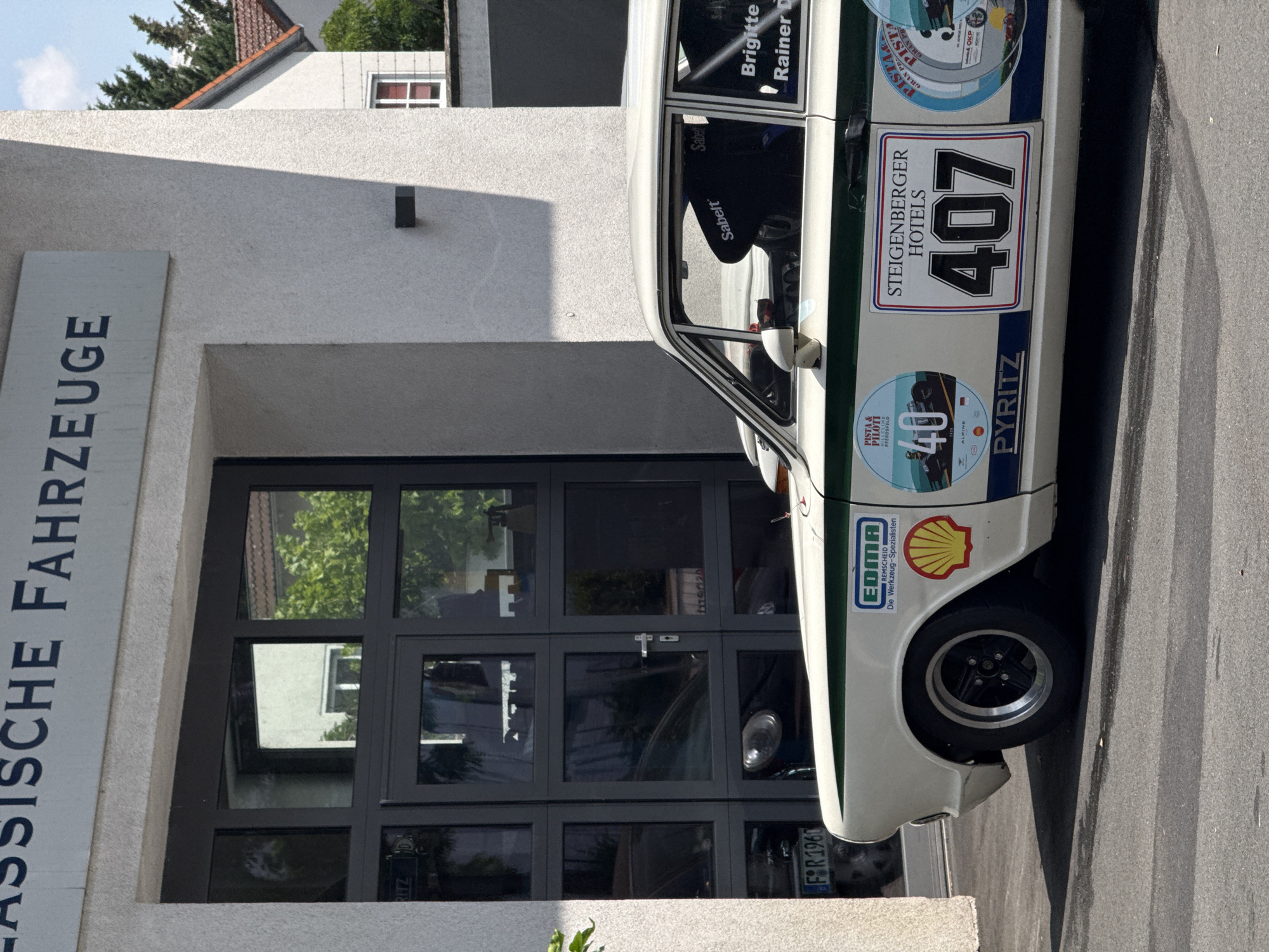 The image depicts a white classic car, possibly a Ford Taunus, with green and blue racing stripes and various stickers on its side, parked in front of a building. The building has a large gray sign above its entrance that reads "KLASSISCHE FAHRZEUGE" in dark blue lettering, which translates to "CLASSIC CARS". The entrance consists of large, dark-framed glass doors. A black racing seat is visible through the driver's side window of the car, with the "Sabelt" logo clearly displayed on it. On the car's door, a sticker for "STEINBERGER HOTELS" with the number "407" is prominent, alongside other circular stickers, one of which reads "Pista & Piloti" and "40". Another sticker indicates "EDMA" with the slogan "Die Werkzeug-Spezialisten" and a Shell logo. The car's license plate is partially visible and reads "FSR 1960". Reflections in the glass doors show the interior of what appears to be a showroom or garage, with other vehicles and possibly car parts visible. The surrounding environment suggests an urban or suburban setting, with a house and trees visible in the background. The car is positioned on an asphalt surface, possibly a street or parking lot, with tire marks visible. The lighting suggests it is daytime, with shadows cast on the ground. The scene is located in Offenbach am Main, Germany, as indicated by the presence of the building with the "Klassische Fahrzeuge" sign, which is a known dealership for classic cars in that city.