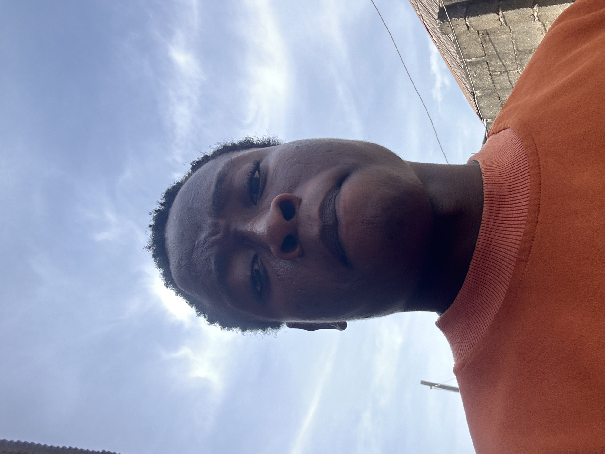 A low-angle perspective photograph captures a person of African descent against a backdrop of a partly cloudy sky and urban elements. The person, positioned close to the camera, fills the frame with their face, which features a dark complexion, short curly hair, a prominent nose, and partially visible eyes. They are wearing an orange-colored sweatshirt or long-sleeved shirt, of which the collar is visible.
The sky, a pale blue, is punctuated by scattered white clouds. In the upper right corner, a portion of a brick wall is visible, possibly part of a building. A thin electrical wire or cable runs diagonally across the sky. The location is within an urban environment, but the specific city is unknown.
