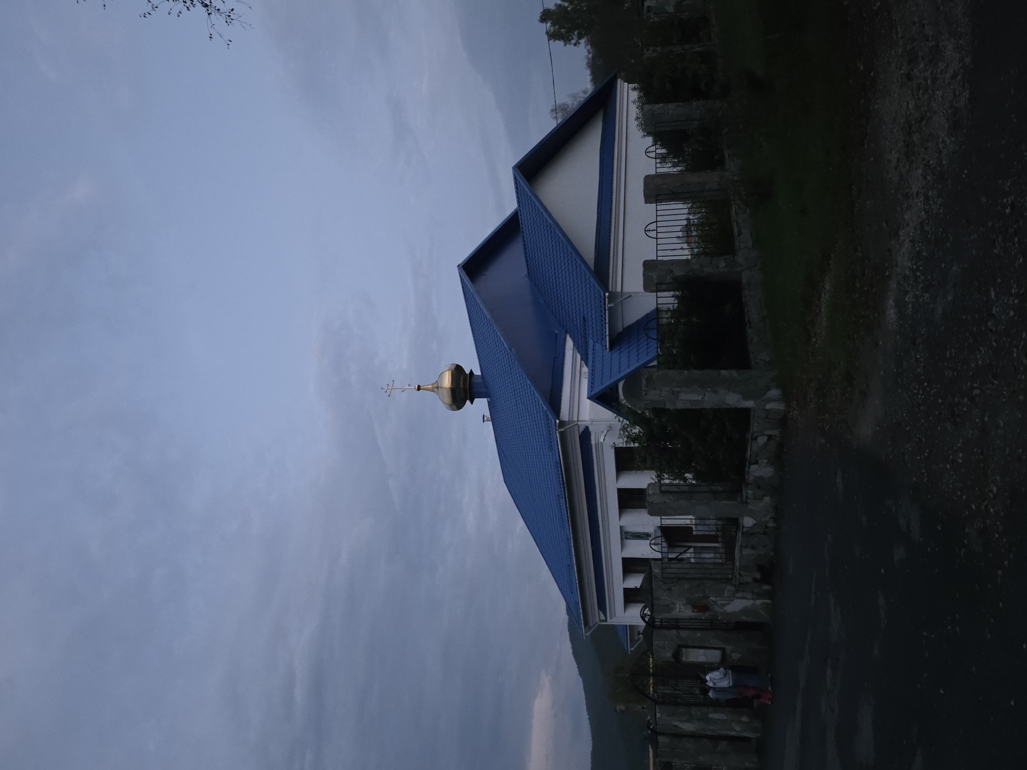 An outdoor, low-angle, eye-level, medium shot captures a church with a blue roof and golden dome in the town of Magnitka, Russia. The sky is overcast with grayish-blue clouds. In the foreground, a dark asphalt road leads to a stone fence surrounding the church. Two people, a woman and a child, stand near the fence. The church is white with a blue roof and a golden dome topped with a cross. Trees and bushes are visible in the background. The overall lighting suggests it is twilight.