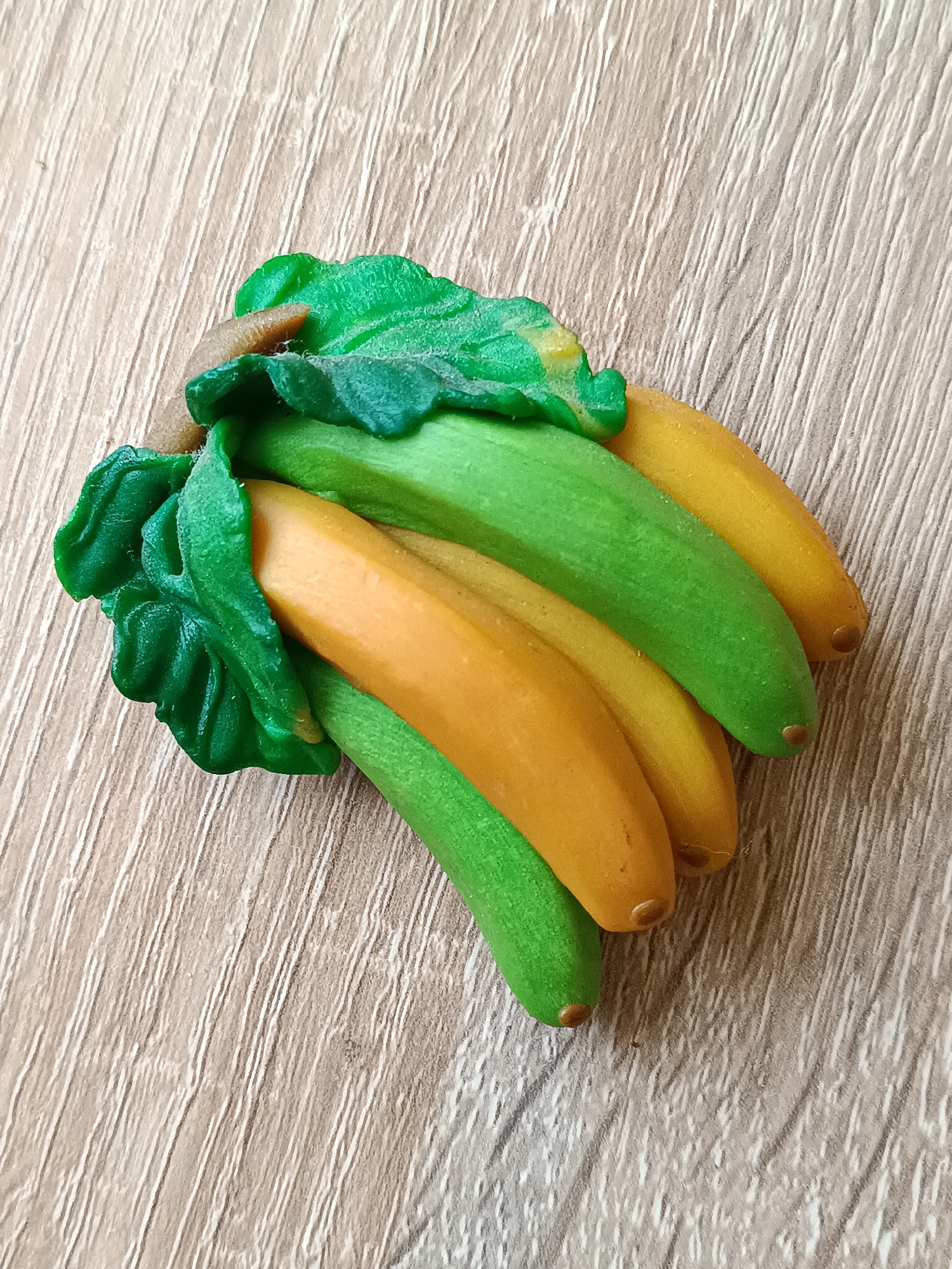 A bunch of artificial bananas, some green and some yellow, are arranged on a light brown wooden surface. The bananas are clustered together, with green leaves at the base. The surface has a wood grain texture. The scene is viewed from a top-down perspective. No people are present, and no action is taking place. The image provides no information about the city of Tebras, Latvia.