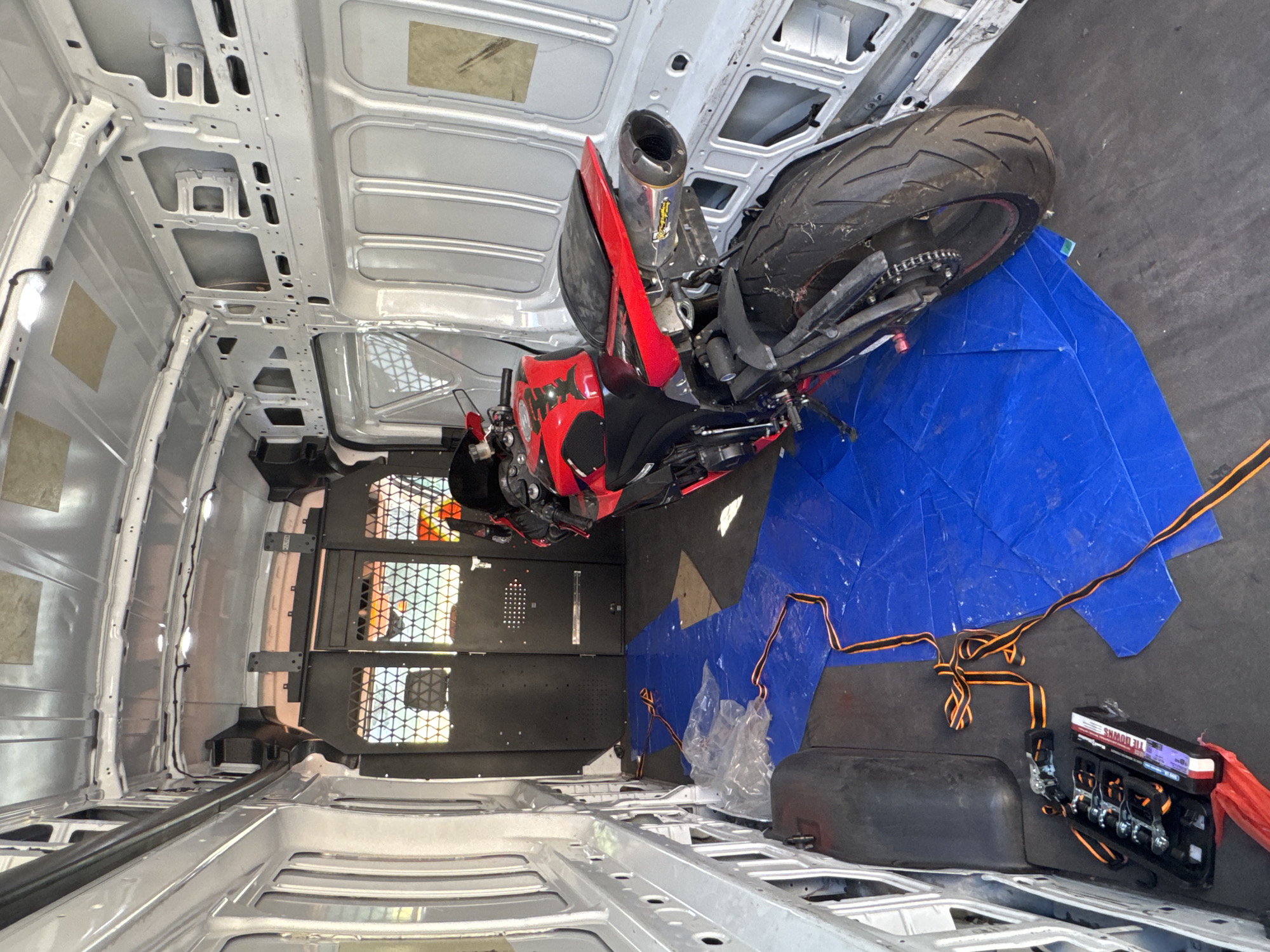 The image depicts the interior of a white cargo van, likely parked in Citrus Heights, California. A red and black sport motorcycle is positioned inside the van, resting on a blue tarp spread across the floor. The tarp is partially covered by clear plastic sheeting and orange and black tie-down straps, suggesting the motorcycle is being prepared for transport. A black case containing tie-down ratchets is visible on the floor to the left of the motorcycle. The van's interior appears utilitarian, with bare metal walls and ceiling, and a caged partition at the rear. Sunlight streams through the rear doors, illuminating the scene.