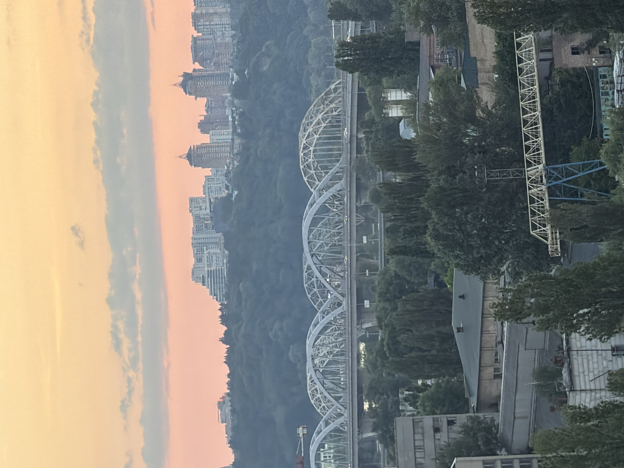 This image shows a city skyline with multiple bridges and buildings. The sky is a gradient of orange and pink, suggesting either sunrise or sunset. In the foreground, there are industrial buildings and trees. In the midground, a large, arched bridge spans across a body of water, with cars visible on its roadway. Further in the background, a dense green hill is topped with a cluster of modern buildings, some of which appear to be high-rise apartments. The overall scene depicts an urban landscape in Kyiv, Ukraine, characterized by a mix of infrastructure and natural elements.