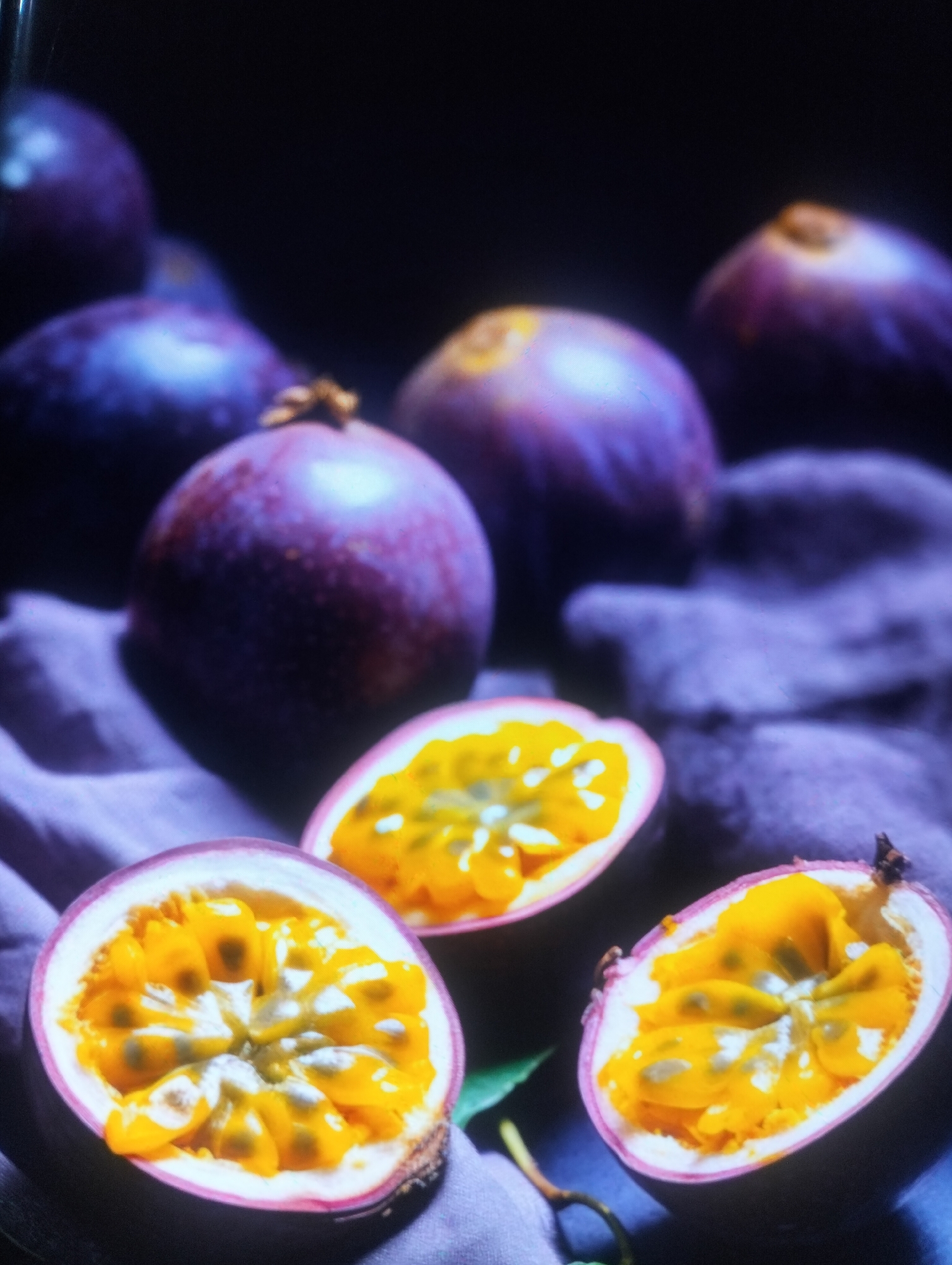 The image depicts a still life arrangement of passion fruits on a dark, textured surface, possibly a table or cloth. Several whole passion fruits, predominantly deep purple with hints of pink, are visible in the background, appearing somewhat out of focus. In the foreground, three passion fruits are cut in half, revealing their vibrant yellow, gelatinous pulp filled with small, dark seeds. These cut halves are arranged in a slightly overlapping manner, with the pulpy interiors facing upwards. A dark purple fabric, possibly a tablecloth or napkin, serves as the base for the fruits, its folds and texture adding depth to the composition. The lighting is dim and moody, casting soft shadows and highlighting the glossy sheen of the fruits. There are no people, animals, or any indication of a city or specific location like Payagaboh, Indonesia present in the image.