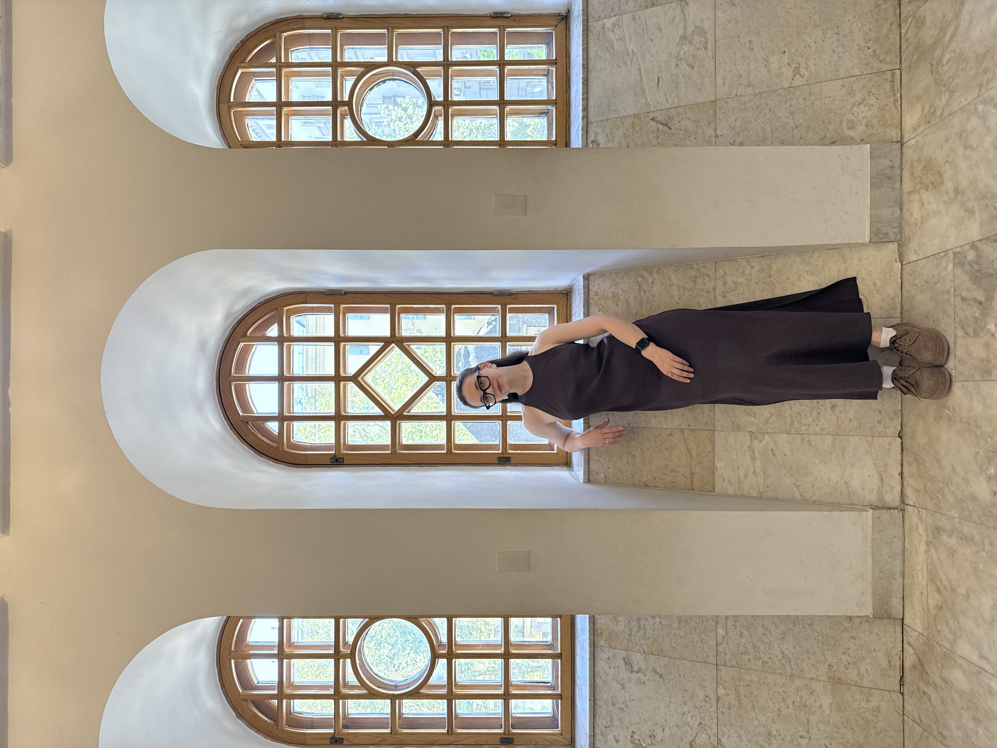 A woman wearing glasses, a dark brown dress, and light brown shoes is standing between a column and a wall, near a large window, inside a building. The building has three arched windows on the wall, each of which has multiple rectangular panes and a circular window in the center. The environment suggests that the location may be in a building in the city of Moscow, Russia. The floor and wall are made of tan marble.