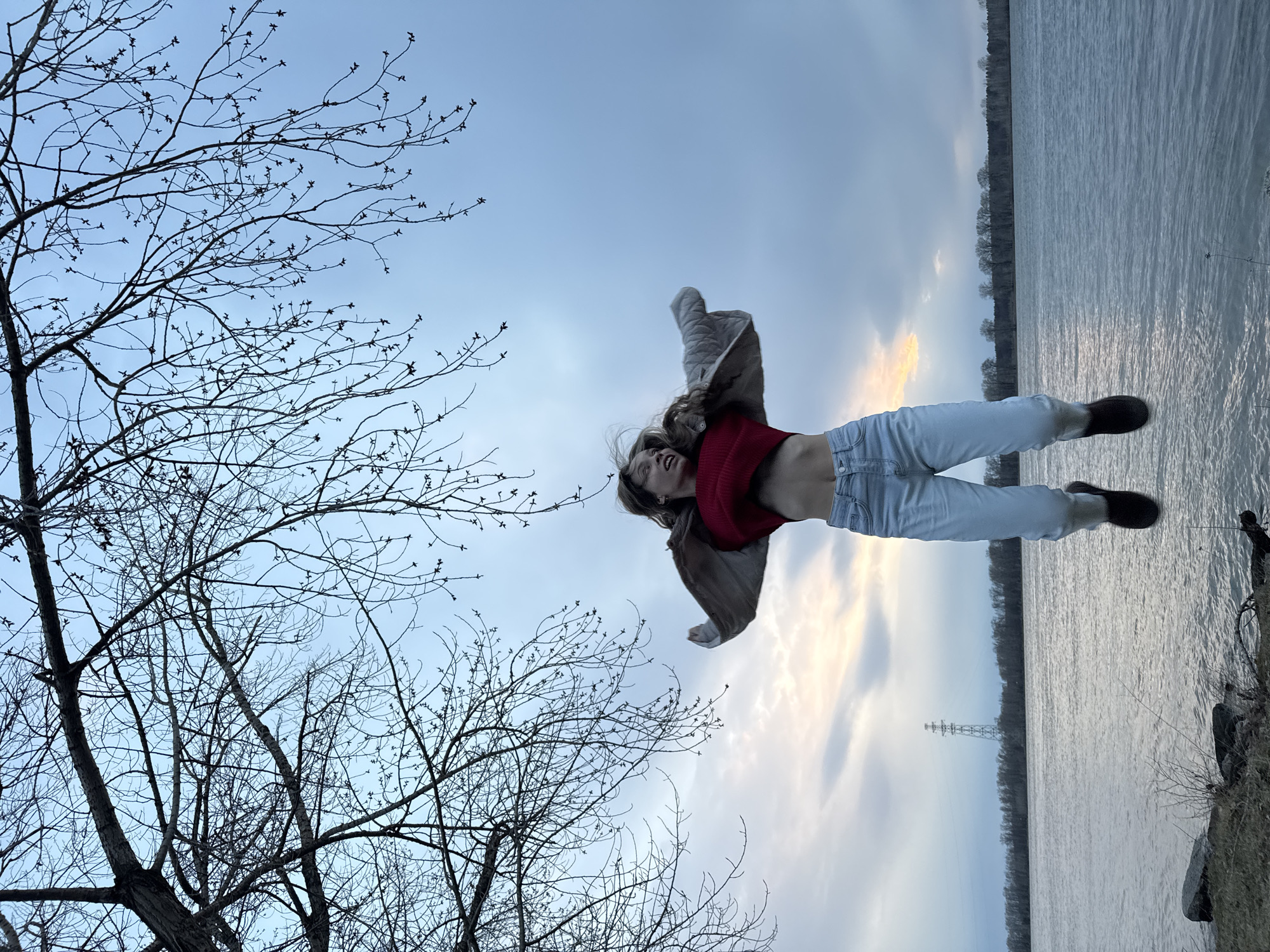 This image, originally rotated, captures a dynamic outdoor scene at dusk or sunset in Chaikovsky, Russia. A young person, appearing to be female, is the central subject, captured mid-air or in a joyful, energetic pose. She is smiling broadly with her head tilted back, and her hair is flying, suggesting movement. She wears a red cropped top, light-colored (likely light-wash denim) jeans, and dark shoes. A light-colored jacket is airborne behind her, further emphasizing the sense of motion.
In the foreground, on the left side of the frame (when correctly oriented), are the stark silhouettes of bare tree branches, showing numerous small buds, indicating early spring or late winter. The background features a wide body of water, likely a river or lake, with visible ripples reflecting the sky. The distant opposite bank is lined with a dense strip of bare trees, and a tall electricity pylon is visible further in the distance. The sky is overcast but displays soft patches of yellow and orange, characteristic of either a sunset or sunrise. The overall atmosphere conveyed is one of joy and freedom. There is no visible text in the image.