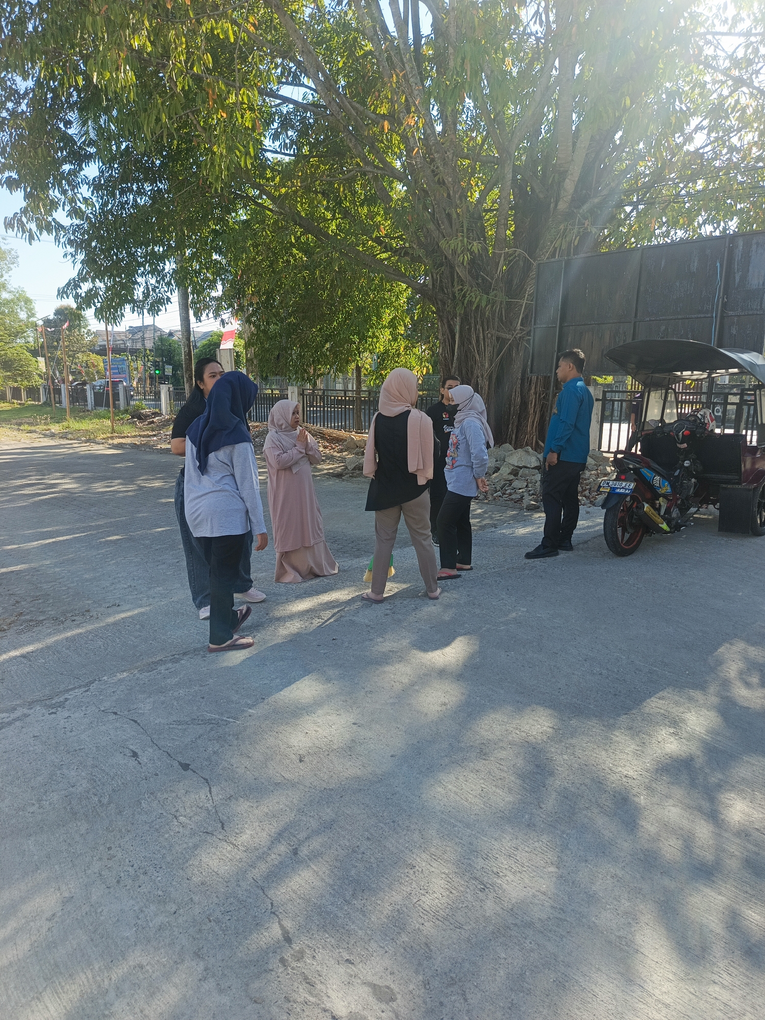 A group of six people are standing on a paved road in Gorontalo, Indonesia, under a large tree. The sun is shining brightly, casting shadows on the ground. The people appear to be conversing. One person in a blue shirt is standing near a small motorized vehicle that resembles a modified motorcycle with a cart attached. The vehicle is parked on the side of the road. Buildings and fences are visible in the background, suggesting an urban or suburban setting. The road surface shows some cracks and is uneven in places.