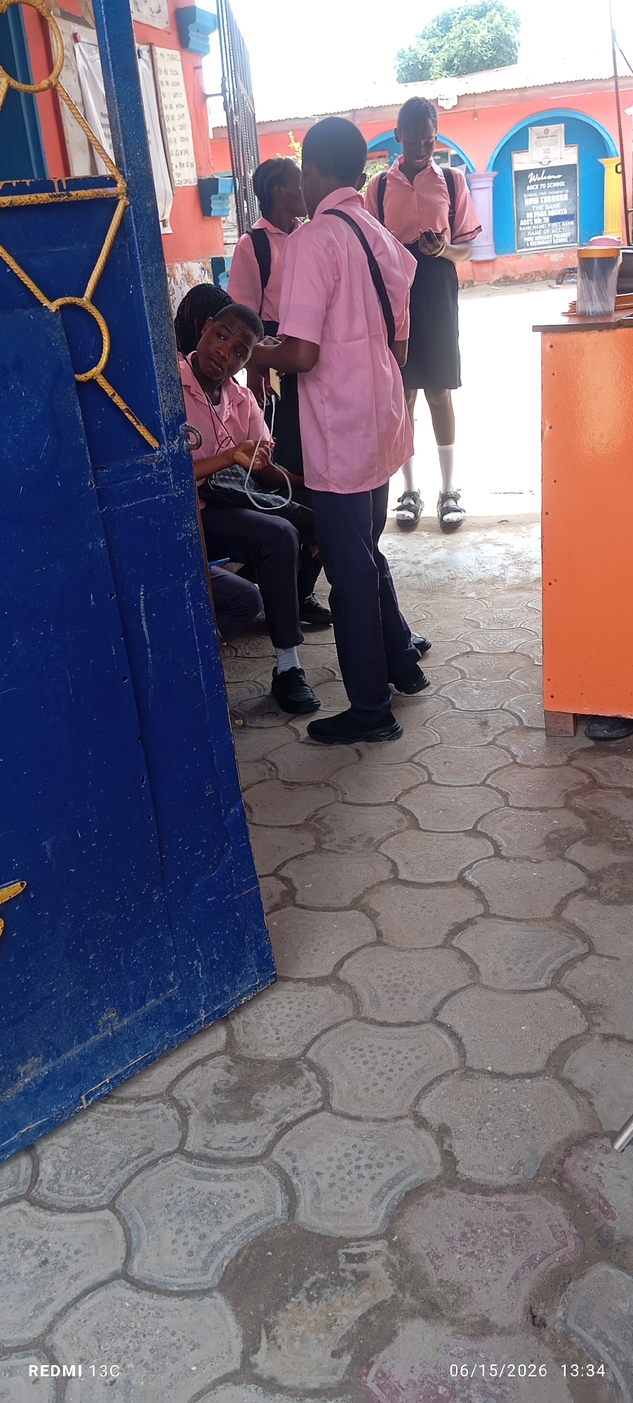 The image shows a group of young people, likely students, standing near a blue gate. They are wearing pink shirts and dark trousers or skirts. One student in the foreground, partially obscured by the gate, is looking towards the camera. Another student in the middle ground is holding a mobile phone. In the background, there is a building with orange and blue accents and what appears to be a welcome sign. The ground is paved with interlocking concrete blocks. The scene suggests an outdoor setting at what could be a school or community center in Lagos, Nigeria, given the context of the REDMI 13C phone in the lower left corner and the date stamp "06/15/2026 13:34" in the lower right.