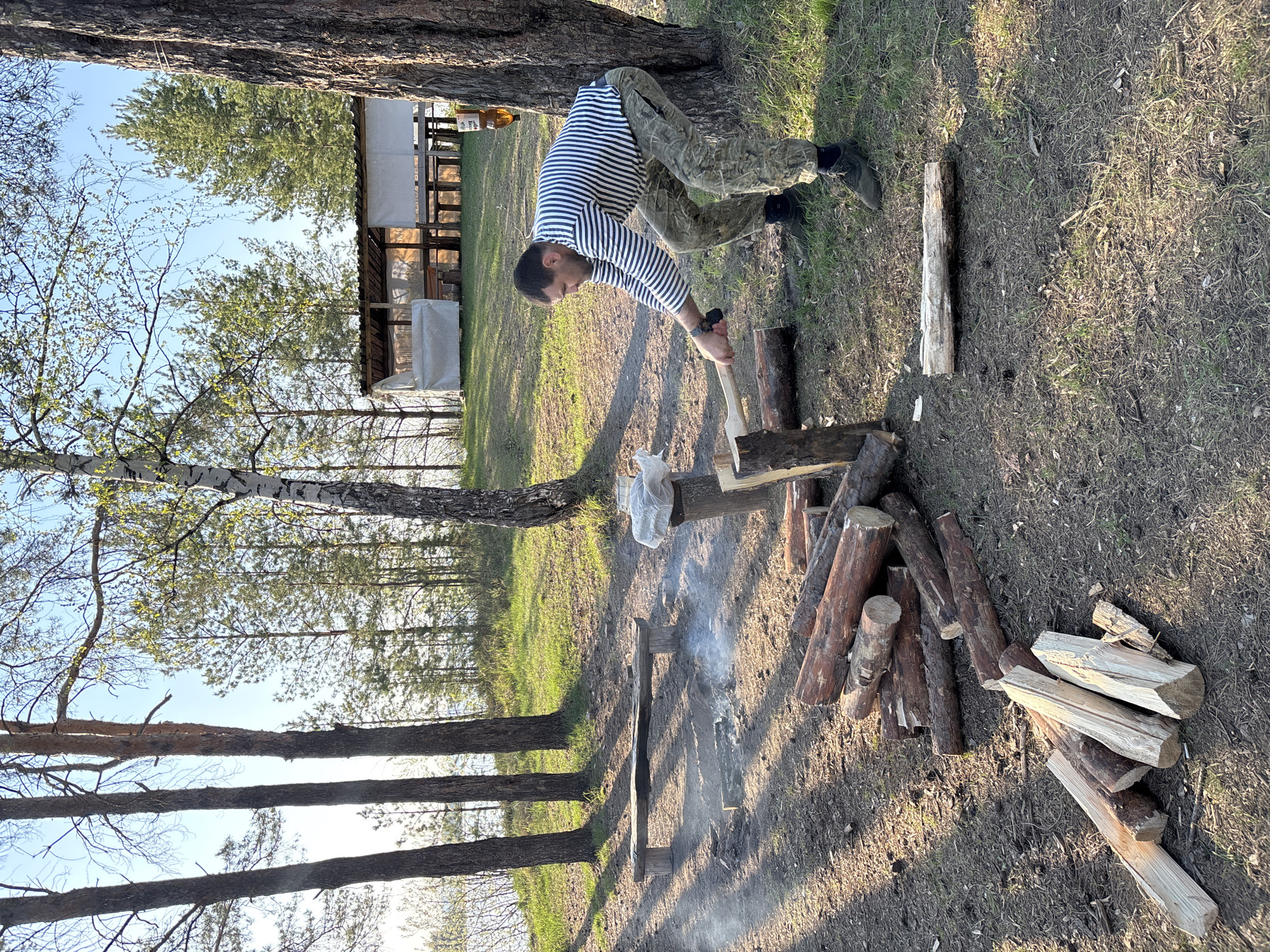 The image shows a man in a striped shirt and camouflage pants chopping wood outdoors. He is using an ax to split a log on top of a stump. A pile of chopped wood and unchopped logs are nearby, along with a small fire pit with smoke rising from it. The setting appears to be a wooded area, possibly in Vekoshinka, Russia, based on the prompt. A building is visible in the background.