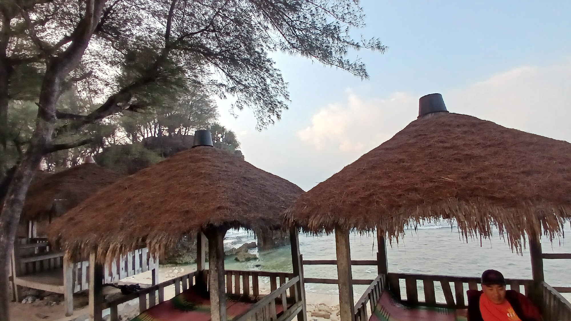 This image shows a beachside scene in Tepus, Indonesia, featuring several thatched-roof gazebos with wooden structures. The gazebos are situated on a sandy area, with the ocean visible in the background. Trees with sparse foliage are present on the left side of the frame, with a rocky outcrop and the sea further back. The sky is overcast with some clouds. One gazebo in the foreground has a dark bucket placed on top of its thatch. A person wearing a dark cap and an orange shirt is seated inside another gazebo on the right, facing away from the viewer. The water appears calm with gentle waves.