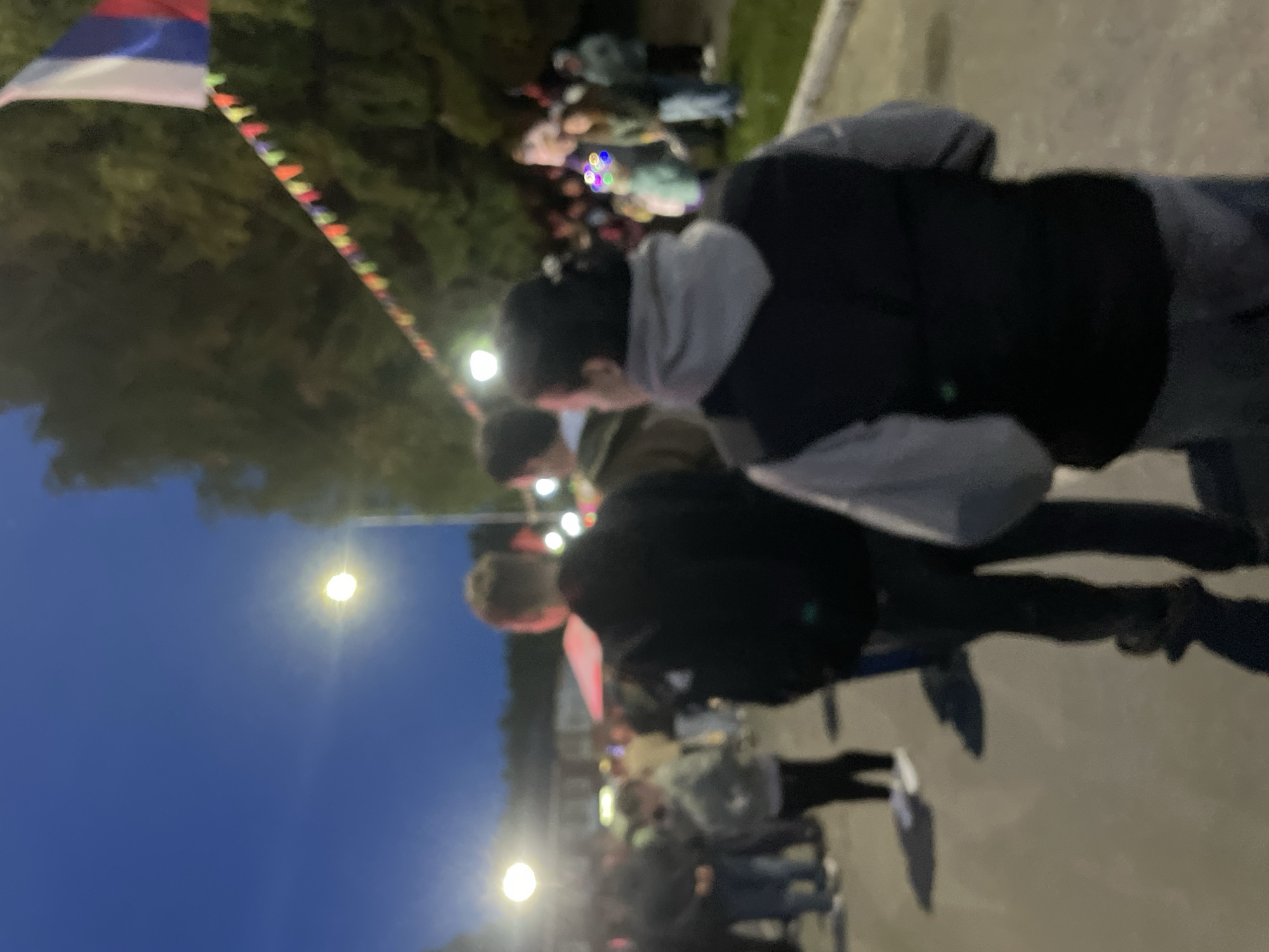 The image appears to be taken at night in Красные Баки, Russia, judging from the Russian flag visible. A small crowd of people is illuminated by streetlights. Three individuals are in the foreground, partially obscuring the view of the crowd. The primary subject seems to be the gathering of people. The environment includes trees and what looks like a paved surface. Colorful flags or decorations are strung along a line.