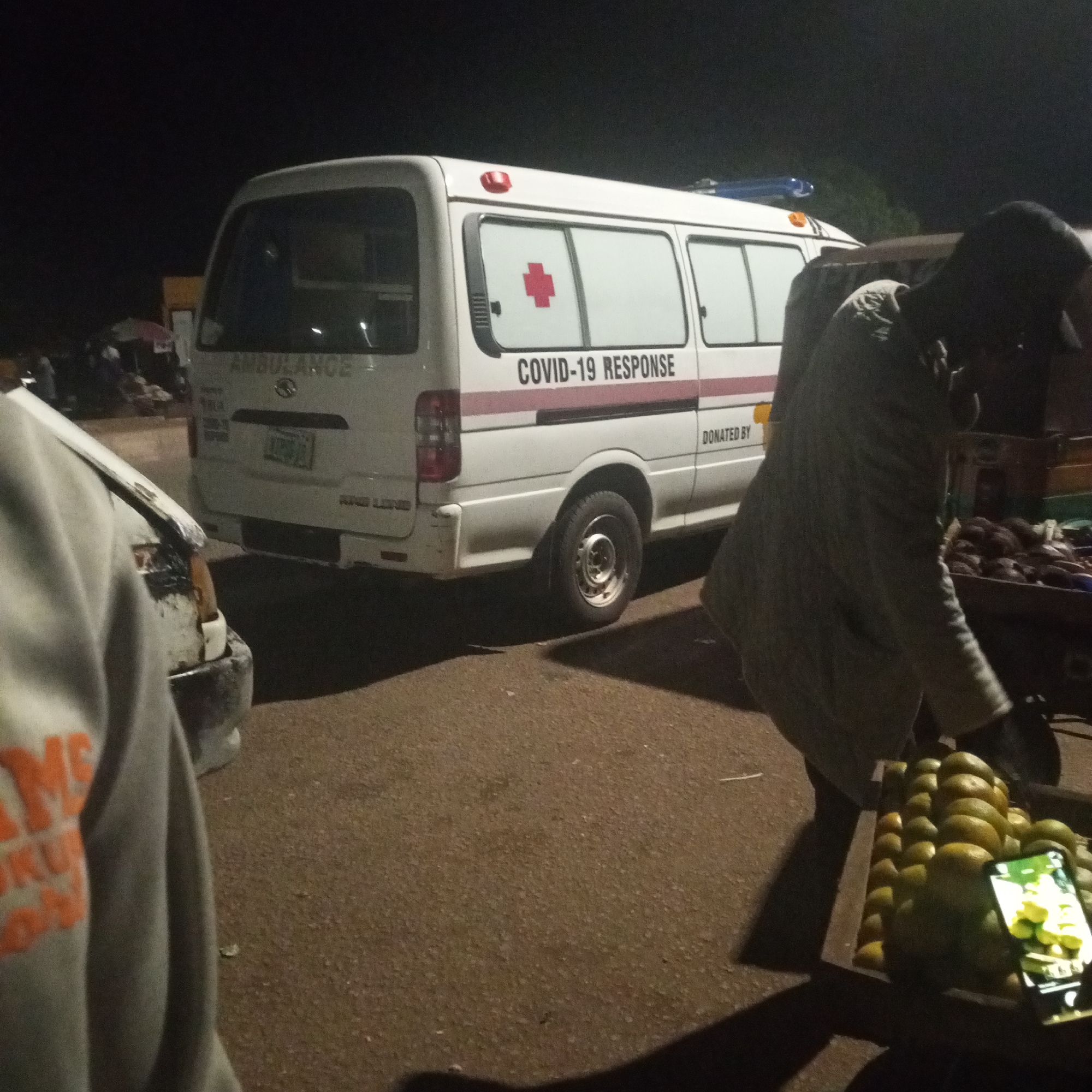 A white ambulance labeled "COVID-19 RESPONSE" is parked on a dirt road at night in Bukuru, Nigeria. The ambulance has a red cross emblem on its side. A person wearing a light-colored jacket stands near the ambulance, appearing to be looking at their phone. In the foreground, a person wearing a gray hoodie with orange lettering is partially visible, and to the right, a wooden crate filled with oranges is on display. The background is dark, with some indistinct shapes suggesting structures and possibly other people or market stalls. The scene appears to be outdoors, possibly a marketplace or a roadside stop.