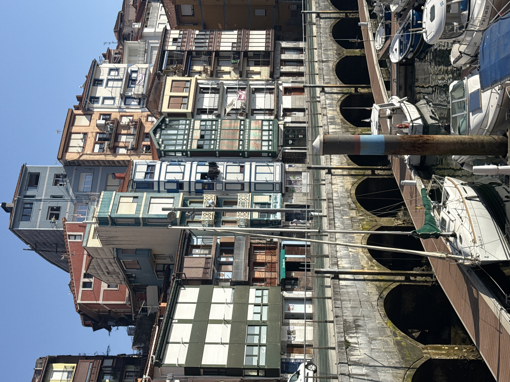 The image depicts a waterfront scene in Bermeo, Spain, featuring a row of colorful buildings lining a harbor. The buildings have diverse architectural styles, with multiple stories, balconies, and prominent windows. A stone bridge with arches spans the water in the foreground, with several boats docked in the harbor below. The boats are of various sizes and types, including a sailboat and several smaller motorboats. The water reflects the buildings and the sky, which is a clear blue. The scene appears calm and sunny, with no people actively engaged in any visible activity.