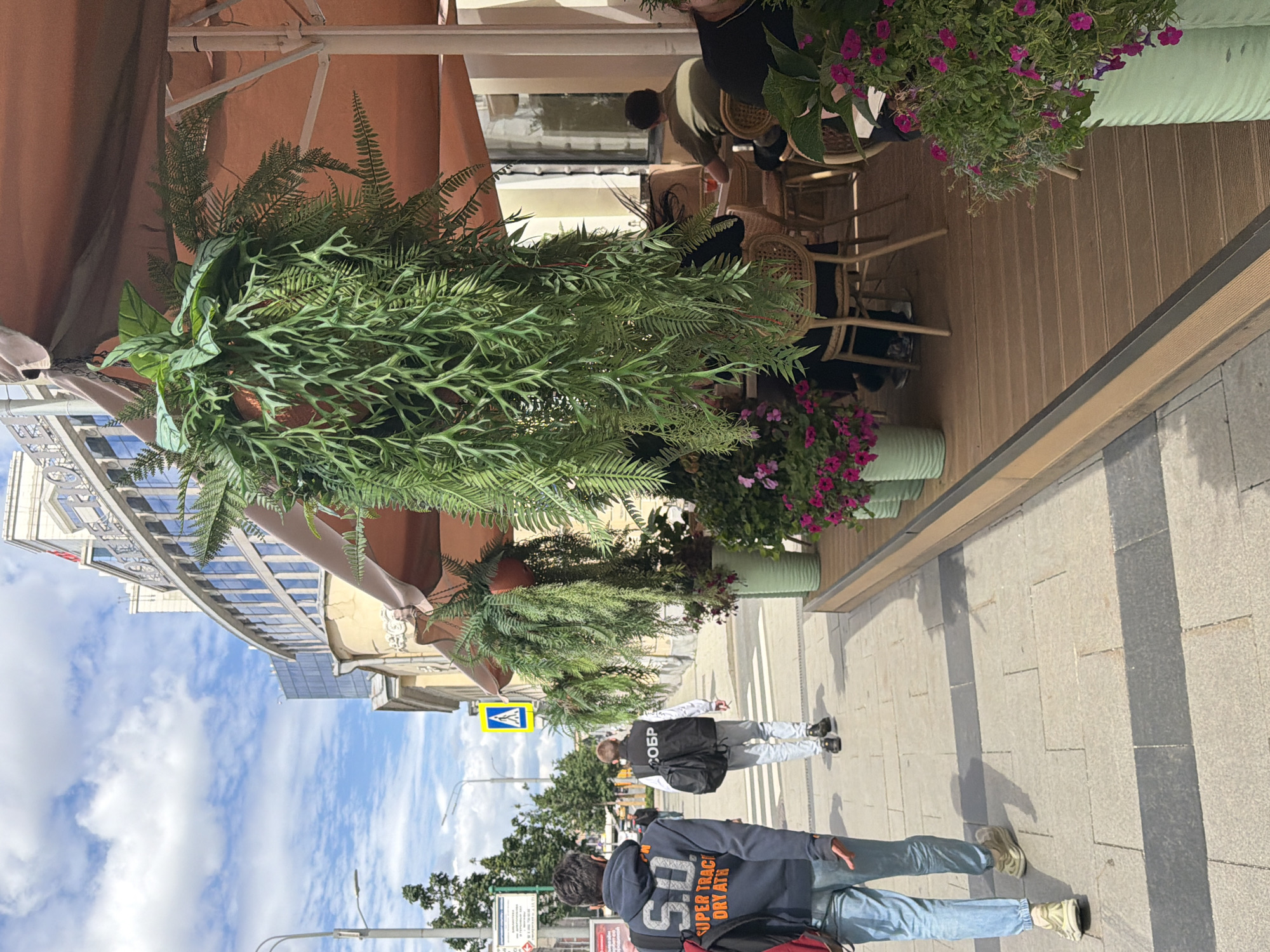The image depicts a street scene in Moscow, Russia. Three individuals are visible, two walking and one appearing to be seated at a cafe. The foreground shows the legs of two people, one wearing a dark hoodie with the text "SUPER TRACK DRY ATH" and the other a light-colored hoodie. They are walking on a paved sidewalk. In the mid-ground, another person in a dark vest with the Cyrillic text "СОБР" (SOBR, a Russian special purpose police unit) is walking away from the camera. The scene includes abundant greenery, with large potted plants and hanging ferns adorning an outdoor cafe area. The cafe itself features wooden decking and wicker chairs. In the background, a multi-story building with a curved glass facade and an older, ornate building are visible, suggesting an urban environment. The sky is blue with scattered white clouds, indicating fair weather. The lighting suggests it is daytime, likely late morning or afternoon, given the shadows. The overall mood appears casual, with people enjoying an outdoor setting.