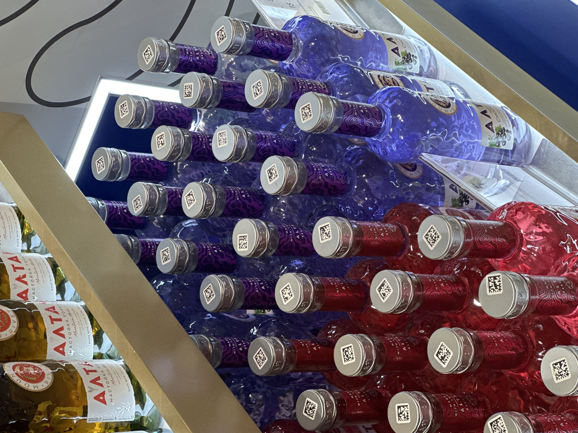 The image shows a display of alcohol bottles in what appears to be a store, likely in a city setting. The bottles are stacked in a visually appealing arrangement, creating a gradient effect with their different colors. Several bottles on the left are green with white labels bearing the name "AATA" and text in Russian. The central section features bottles in shades of purple and blue, while the lower-right section displays bottles in red. The caps of the bottles are silver and contain QR codes. The display includes a gold-colored metal structure, and the background features other architectural elements suggestive of a retail environment.