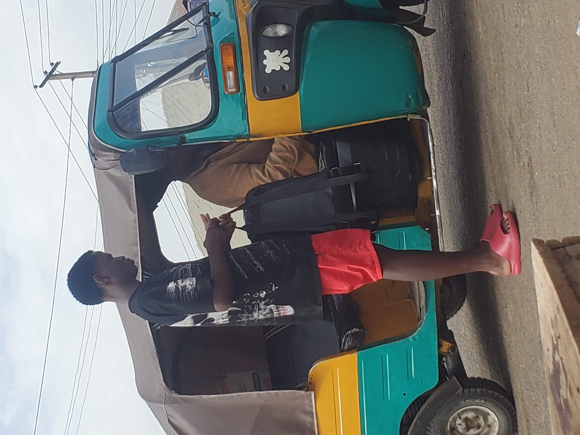 A young Black male, wearing a black t-shirt with a graphic design and red shorts, stands with his back to the camera, looking up and to his left. His right arm is raised, and his hand appears to be making a gesture. He is wearing pink slides. He is standing next to a teal and yellow auto-rickshaw, or "keke," which is common in Nigeria. A person in a brown sweater is visible inside the keke. The keke has a metal frame and appears to be on a paved road. The sky is bright and overcast, suggesting it is daytime. The scene is likely in Gwong, Nigeria, given the presence of the keke and the general setting.