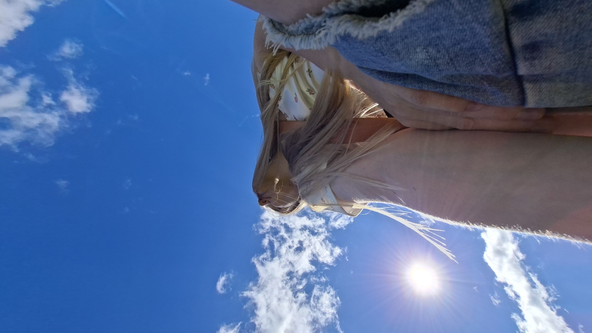The media file shows a low-angle shot of a person's legs and torso against a bright blue sky with scattered clouds. The person appears to be wearing a denim garment, possibly shorts or a skirt, with a frayed hem, and a light-colored top with a floral pattern. Their long blonde hair is visible, falling down and obscuring some of the details. The sun is shining brightly, creating lens flare and illuminating the scene. The perspective suggests the photo was taken from below, looking upwards. There are no visible text elements. The overall impression is one of a warm, sunny day, possibly outdoors in a natural setting. Based on the provided location context, this scene is likely in Bolshoy Bukor, Russia.