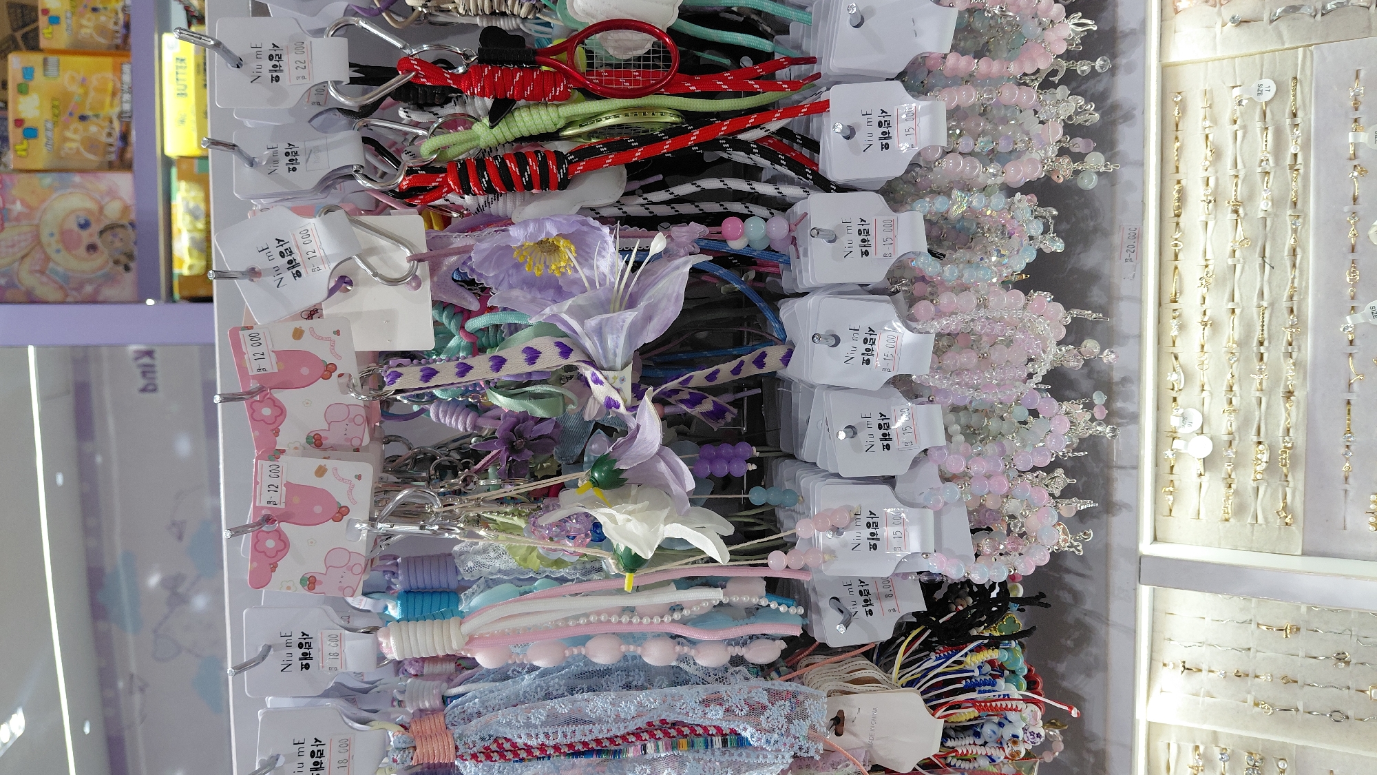 The image shows a close-up view of a retail display of accessories, primarily hair ties, keychains, and bracelets. The items are arranged on several shelves and hanging racks. Many of the accessories are pastel-colored and feature decorative elements like beads, ribbons, and fabric flowers. Some items have price tags attached, indicating prices such as "12,000," "18,000," "15,000," and "22,000." In the background, there are shelves with packaged goods and other displays, suggesting a store environment. The lighting is bright and even, illuminating the products. The scene appears to be within a commercial establishment, possibly a novelty store or a boutique. There are no people visible in the image. The overall impression is one of a colorful and well-organized merchandise display. This image cannot be localized to Purwokerto, Indonesia as there is no information within the image to suggest this location.